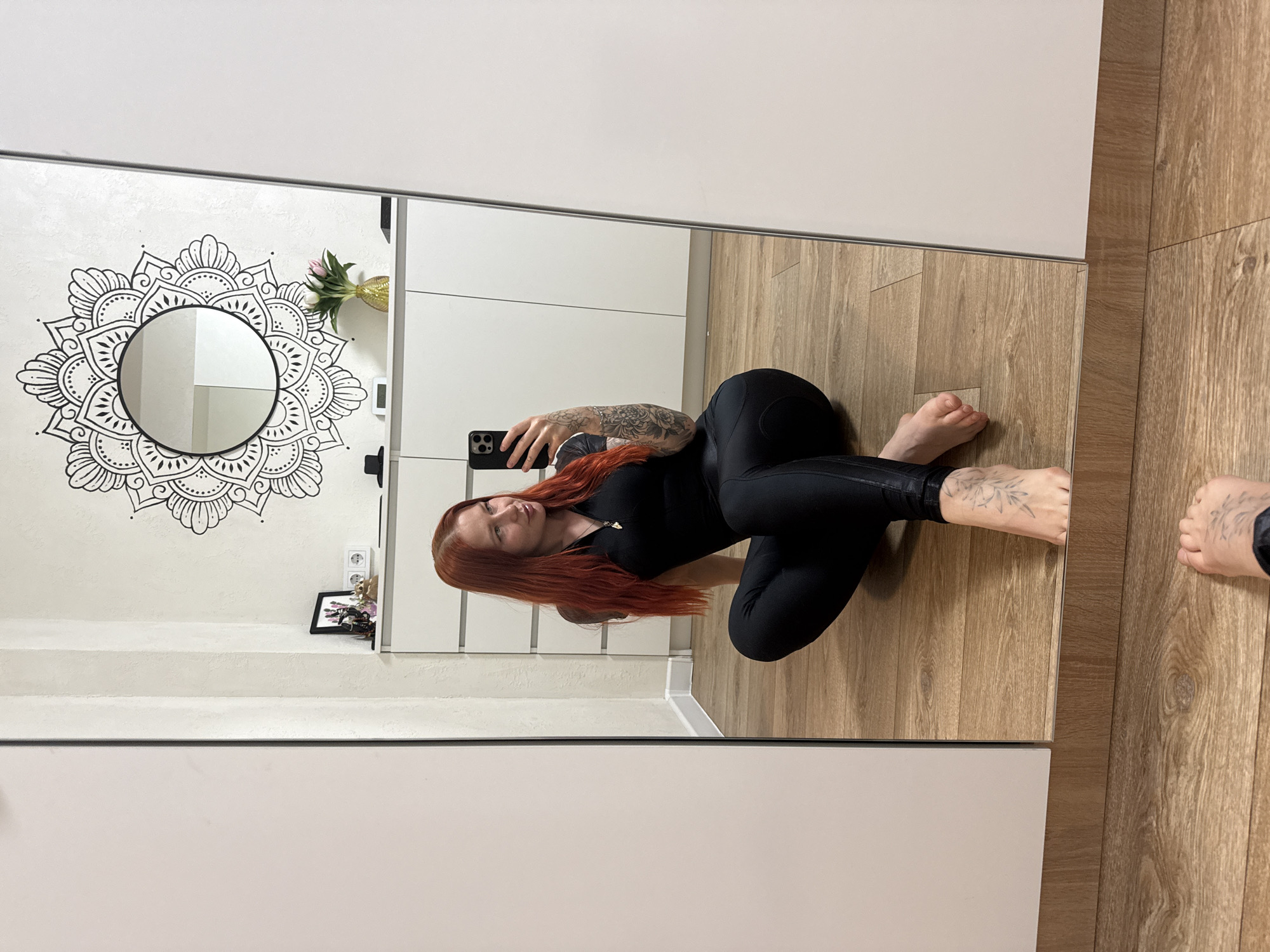 A mirror reflects a woman with red hair wearing a black bodysuit. She is holding a phone and taking a selfie. The reflection shows a wall with a mandala drawing, flowers in a vase, and a picture frame. The floor is wood. No specific location in Rostov-on-Don, Russia, is discernable.