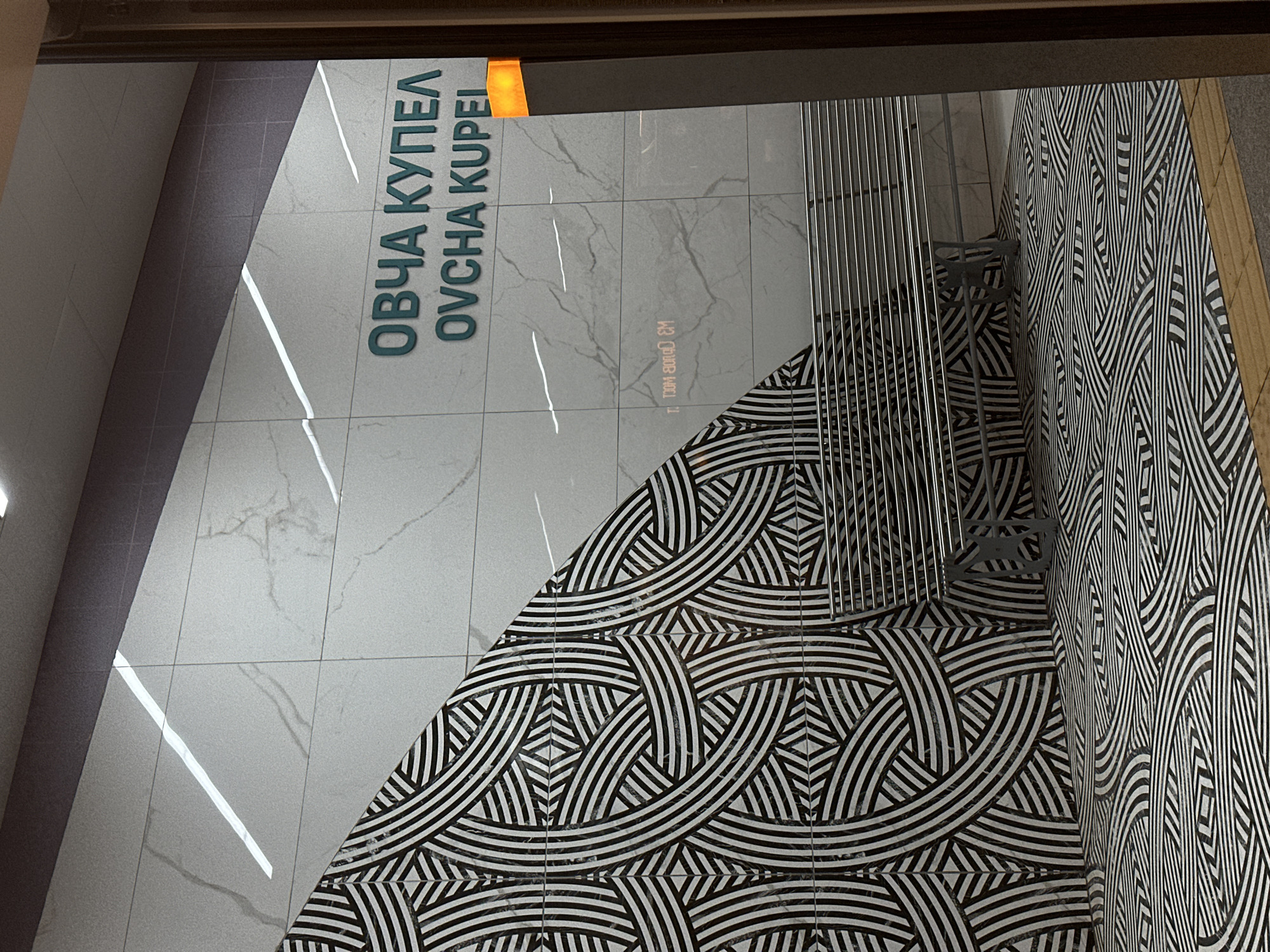 This indoor shot captures a section of the Ovcha Kupel metro station in Sofia, Bulgaria. The image reveals a combination of architectural and design elements, including large green text reading "ОВЧА КУПЕЛ OVCHA KUPEL", which is the name of the station. The walls are tiled with large white-gray tiles, some with marble-like veins. An area of the floor and wall are decorated with an intricate black and white pattern, comprised of curved lines and geometric shapes. A metal barrier or rail is also visible, with an obscured person or object behind the rail. The overall environment suggests a public transportation facility, with a focus on modern design and typography.