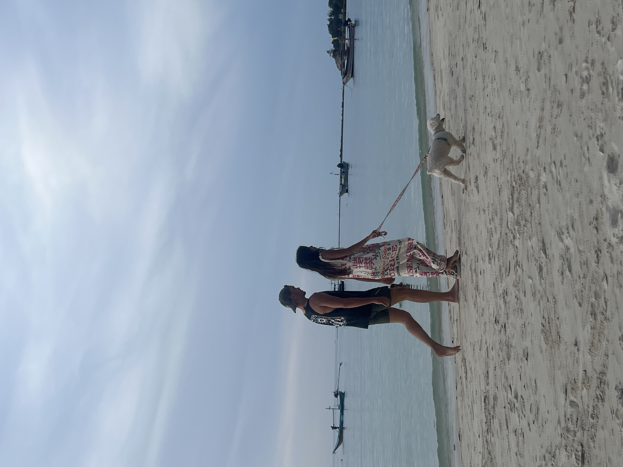 Two people, a male and a female, are walking their light-colored dog along a sandy beach in the Ko Pha-ngan District, Thailand. The male, on the left, is wearing a black tank top, dark shorts, and a baseball cap. The female, on the right, is wearing a long, patterned dress. The dog is on a leash held by the female, and it is walking ahead of them. The ocean is calm and blue, with several small boats visible in the distance. The sky is overcast and a soft gray. The sand is light in color and appears textured with small pebbles and footprints. The image captures a peaceful moment of a couple enjoying a walk with their dog by the sea.