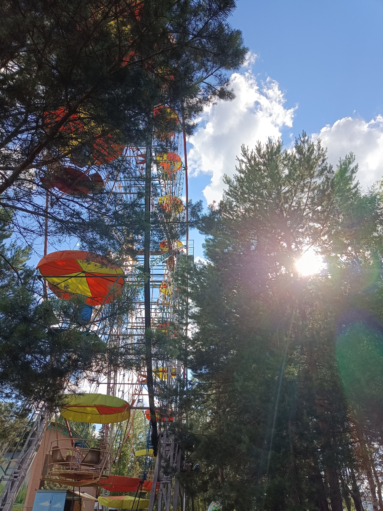 This is a medium-angle, exterior shot taken in the city of Chaikovsky, Russia, showing a Ferris wheel partially obscured by evergreen trees. The Ferris wheel has a rustic appearance, with a metal structure and gondolas painted in bright colors, including yellow, orange, and red. One gondola is prominently visible in the foreground, its umbrella-like canopy casting a shadow. The sky is blue with scattered white clouds, and the sun is shining through the trees, creating a lens flare effect. The overall impression is that of an amusement park or fairground in a natural, wooded setting.