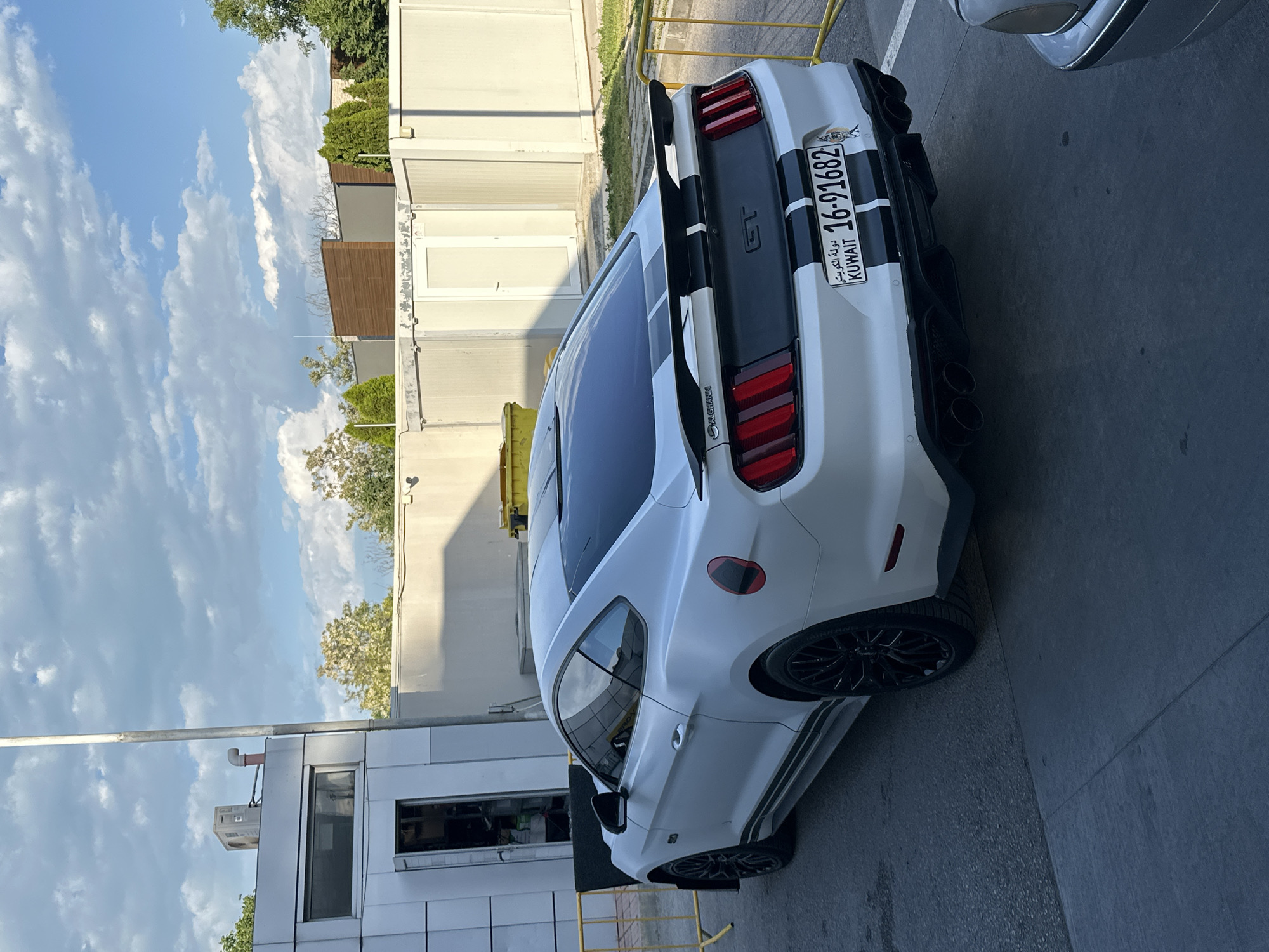 The image depicts a white Ford Mustang GT parked on an asphalt surface. The car features black racing stripes, a rear spoiler, and quad exhaust pipes. The license plate reads "16-91682" and indicates it's from Kuwait. The background includes a building with a modern, industrial design, possibly an office or commercial structure. The sky is visible in the upper left corner, displaying scattered clouds. The location is not Varna, Bulgaria.