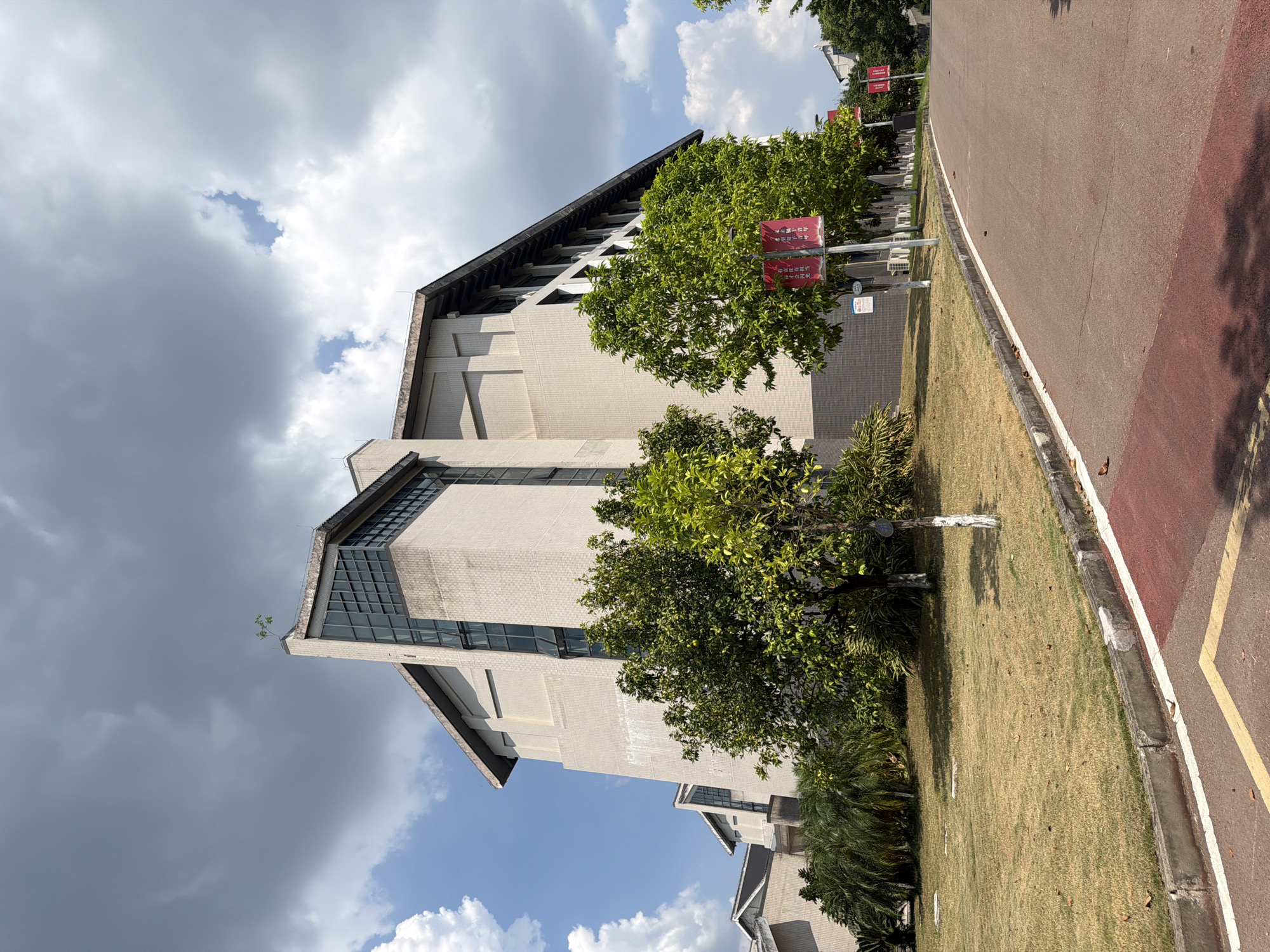 This is an eye-level, outdoor, medium shot of a modern, angular, light-colored brick building with dark windows, located in Jiangjin, China. The building has a prominent, steeply pitched roof with a dark overhang. A small tree grows from the very top edge of the building's corner. The building is surrounded by a well-maintained lawn and several green trees with some bearing what appear to be fruit. Red banners with Chinese text are displayed on poles in front of the building, and a paved road with red and white markings curves away from the viewer in the foreground. The sky is partially cloudy, with patches of blue sky visible through dark grey clouds. The overall impression is one of a public building or institution in a well-kept, possibly academic, setting.