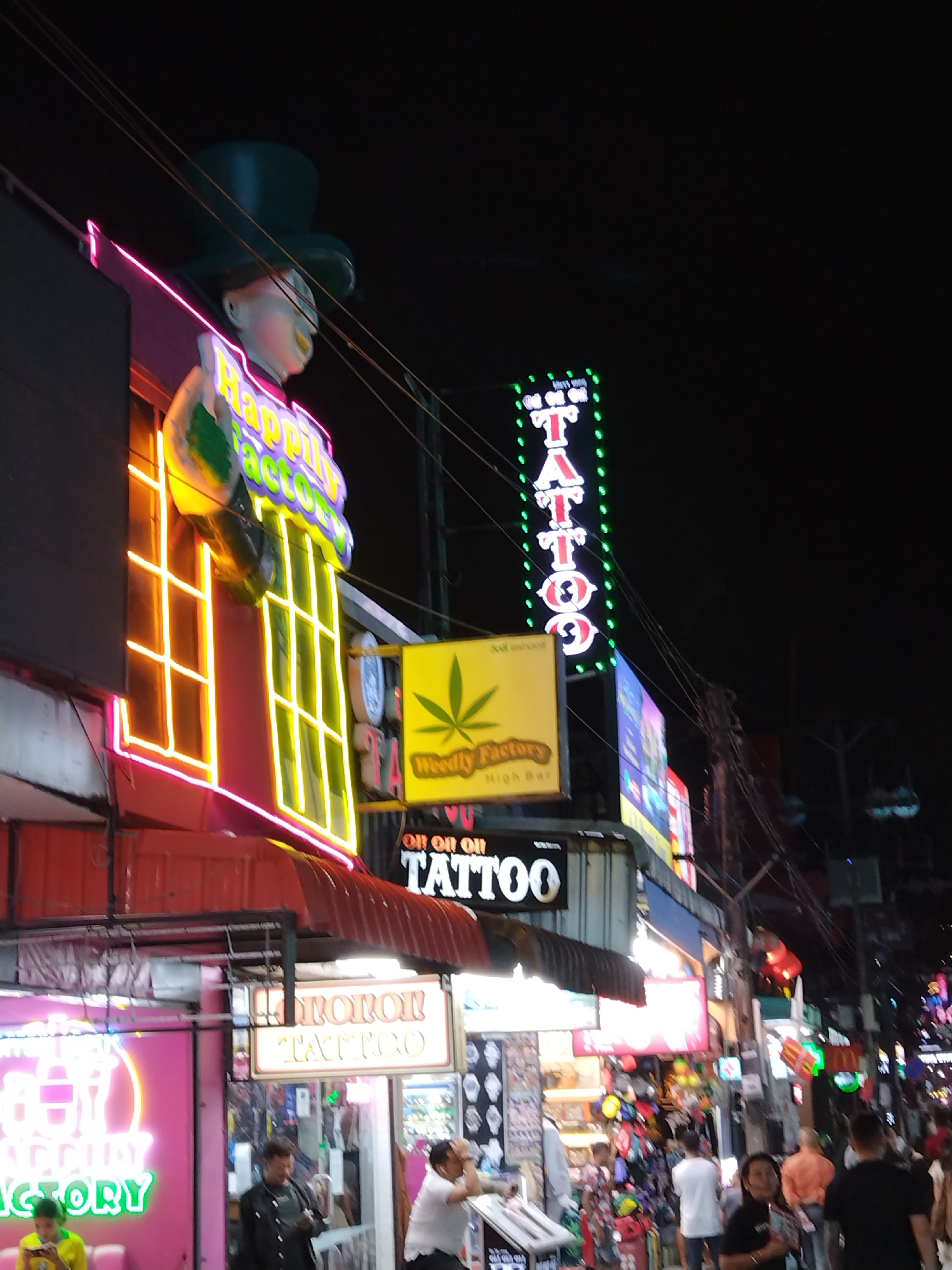 The image depicts a street scene in Patong, Thailand, at night, characterized by vibrant neon signage and a bustling atmosphere. Dominating the left side of the frame is a multi-story building adorned with bright, yellow neon lights forming a window-like pattern, outlined by pink neon. Above this, a large, whimsical statue of a head wearing a dark green top hat is visible. A neon sign reading "Happy Factory" in colorful letters is also prominent on this building.
To the right of this structure, a tall, vertical neon sign flashes the word "TATTOO" in white letters against a green background, bordered by green dots. Below this, a yellow rectangular sign displays a green cannabis leaf symbol and text that appears to say "Weedly Factory" and "High Bar." Further to the right, other neon signs and illuminated advertisements are visible, contributing to the vibrant, commercial ambiance of the street.
The street itself is crowded with people, suggesting a lively nightlife or commercial area. Individuals are seen walking, standing, and interacting. Some are captured in close-up, revealing their expressions and clothing. The lighting from the signs creates a dynamic and somewhat diffused illumination across the scene. Electrical wires and utility poles are visible, indicative of an urban environment. The overall impression is one of a busy entertainment district with a focus on nightlife and potentially adult-oriented businesses, as suggested by the "Tattoo" and "Weedly Factory" signage.