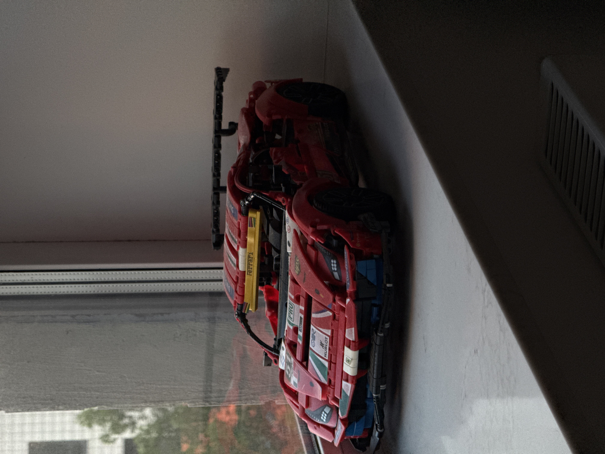 A red Lego Technic Ferrari model car is positioned on a white windowsill. The car is detailed with various stickers, including "Ferrari," "Hublot," and racing numbers. In the background, through a window, a blurred image of a building and trees can be seen, suggesting an urban environment. The windowsill is illuminated by natural light coming from the window, casting shadows on the car and the surface it rests on. The scene is located in the city of Kommunarka, Russia.