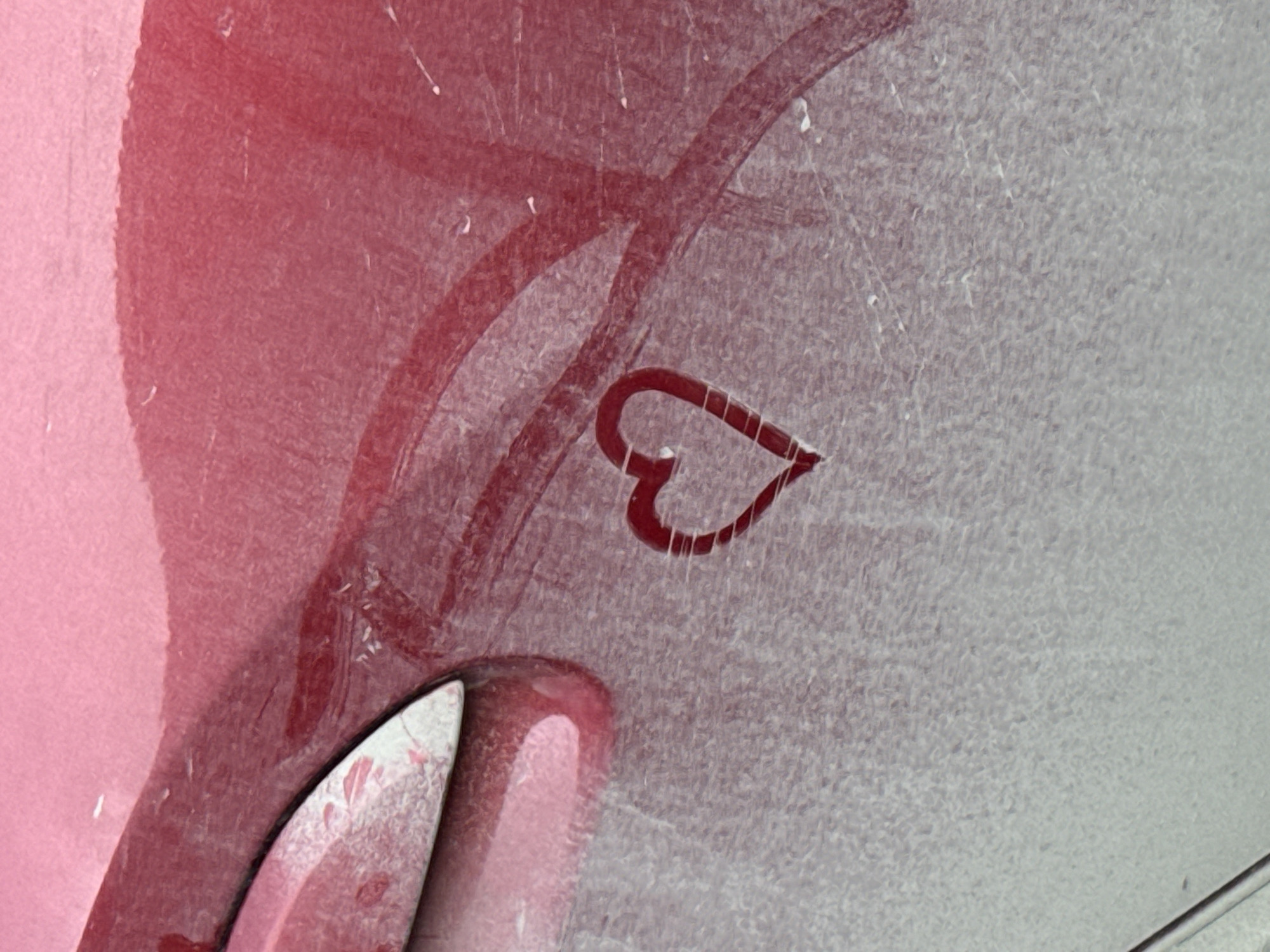 The image shows a close-up of a surface, likely a car, covered in dust. There is a red heart shape drawn in the dust, as well as other red markings or streaks. The background is a mix of light gray and red. There are no people visible in the image. There are no indications of this image being in Izhevsk, Russia.