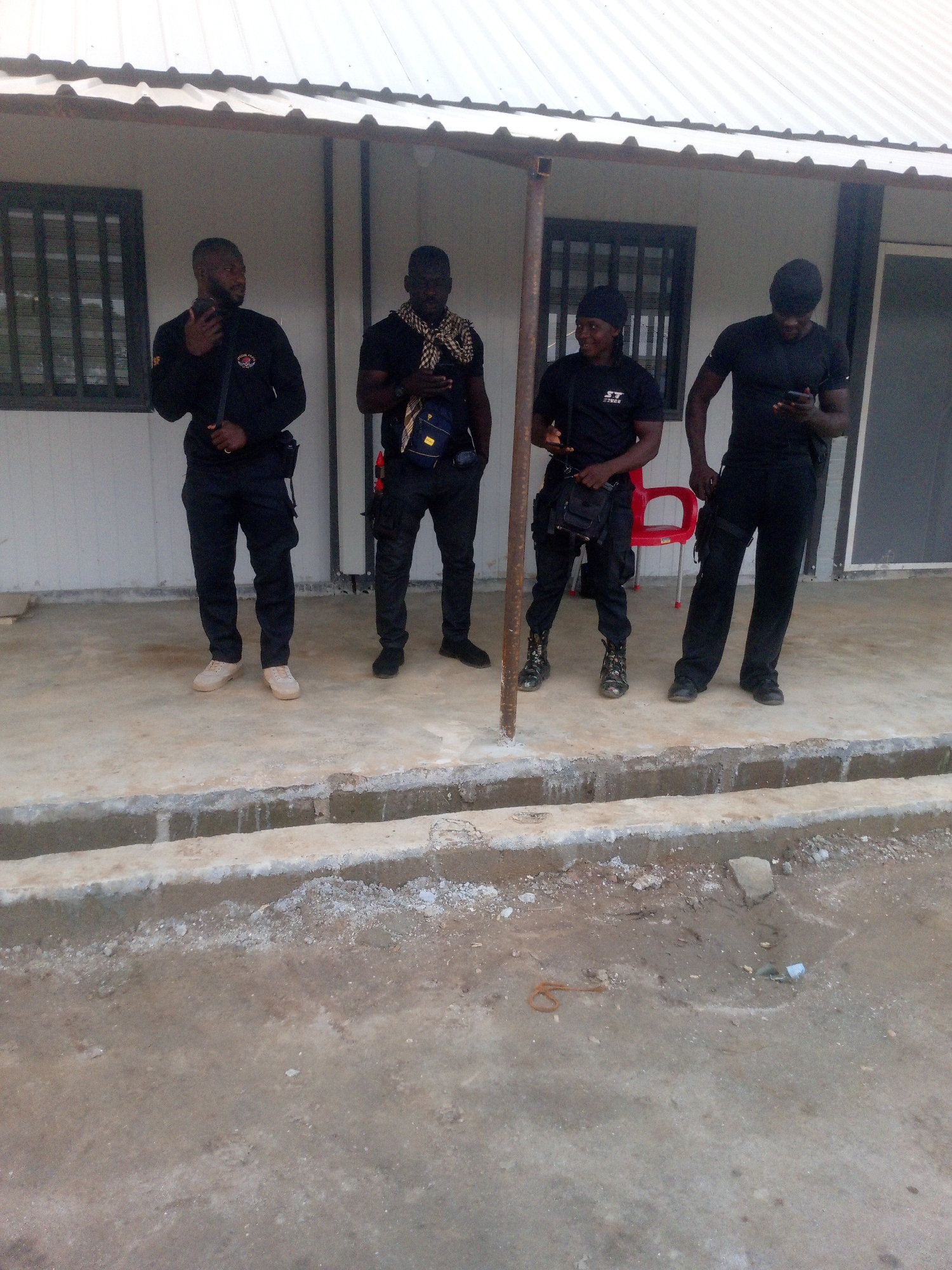 Four individuals, appearing to be men, stand on a concrete outdoor area in front of a white building with a corrugated metal roof. The building has dark-framed windows. All four individuals are dressed in dark clothing, with at least two wearing black t-shirts and black pants. One individual on the far left wears a black long-sleeved shirt and holds a dark object in his right hand. The second individual from the left wears a black t-shirt and a patterned scarf draped around his neck. He has a blue bag slung across his chest. The third individual from the left wears a black t-shirt with a logo on the front and black pants, with a dark bag hanging from his shoulder. He is looking to his left. The individual on the far right wears a black t-shirt and black pants, and is looking down at a dark object in his hands, possibly a mobile phone. A red plastic chair is visible behind the third and fourth individuals. The ground in front of them is concrete with some dirt and debris. The area appears to be outdoors, potentially in a semi-urban or industrial setting, consistent with a location in Indo, Nigeria.