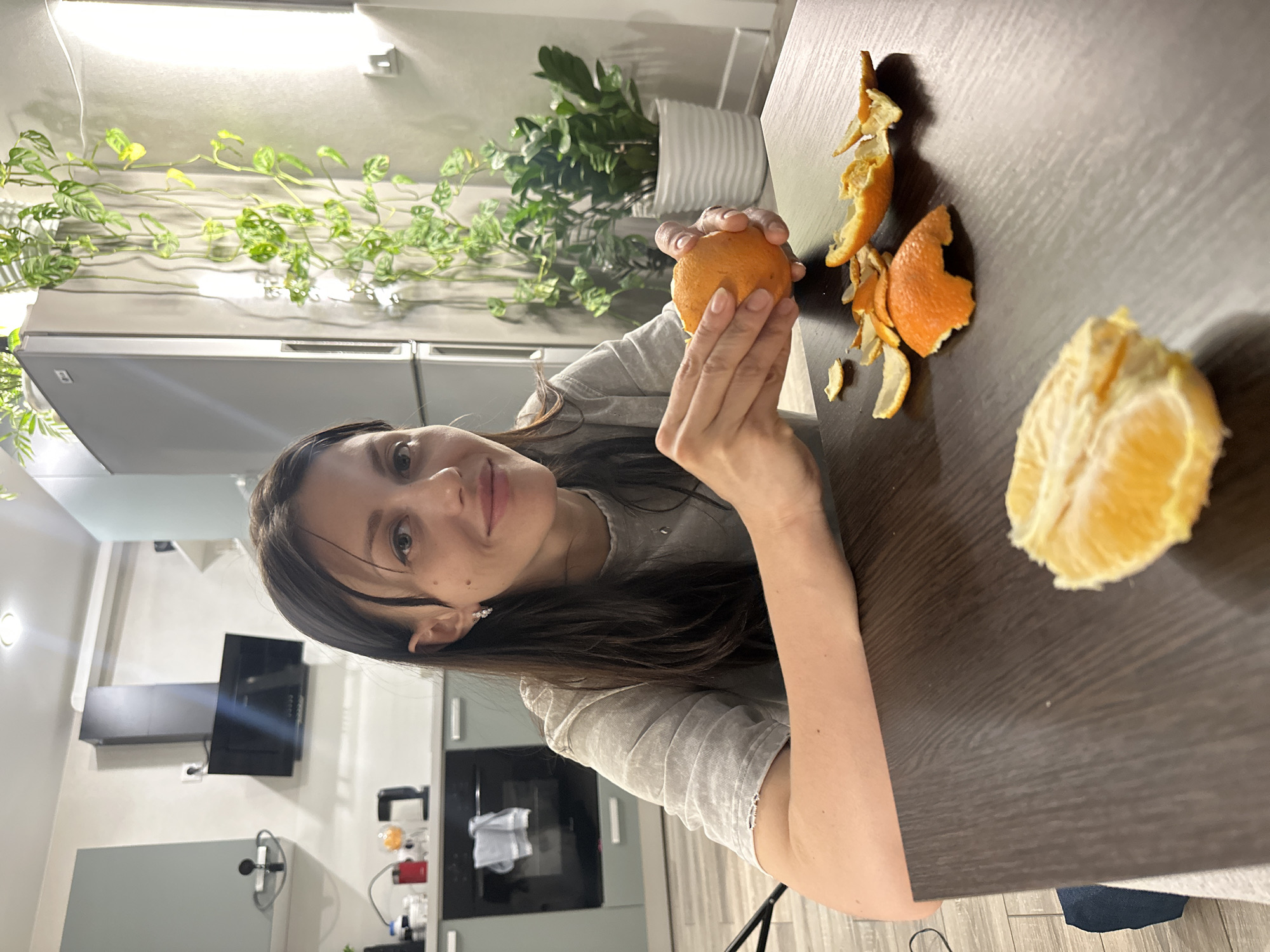 This image depicts a woman peeling an orange in a modern indoor setting. The central subject is a young to middle-aged woman with long, dark hair, wearing a light-colored short-sleeved t-shirt. She has a neutral to slightly pleasant expression and is looking directly at the camera.
She is seated at a dark brown wooden table, holding an unpeeled orange in her left hand, and appears to be in the process of peeling it with her right hand. On the table to her right are discarded pieces of orange peel and a partially peeled orange segment, suggesting the activity is ongoing.
The background reveals a contemporary kitchen or dining area. To the left, a variegated leafy plant (possibly a type of Monstera) trails along a shelf or wall, and another larger potted plant with dense green foliage is visible in a white ribbed planter. The kitchen features sleek, light-toned cabinets, a dark range hood, and other modern appliances.
The lighting appears to be artificial, typical of an indoor home environment. The overall atmosphere is casual and relaxed. There is no visible text in the image. The setting is consistent with a modern residential interior, with the provided context indicating Murino, Russia.