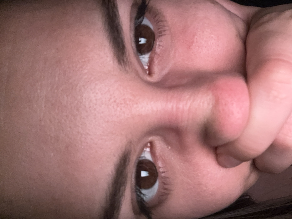A close-up, high-resolution image depicts the upper portion of a human face, spanning from the forehead to the lower nose and mouth, located within an undiscernible indoor environment in Sofia, Bulgaria. The subject presents with dark brown irises, visible dark eyebrows, and light-toned skin exhibiting a reddish cast, possibly due to ambient lighting. A human hand, consistent with the subject's skin tone, is positioned centrally, covering the mouth and the lower portion of the nose. Fingers are visible pressed against the skin. Both pupils display distinct, rectangular reflections of an unknown light source. The immediate background is uniformly dark, providing no additional contextual information.