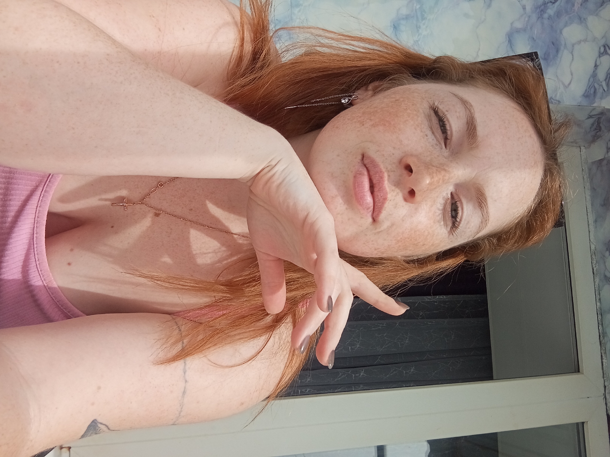 A fair-skinned, auburn-haired person is reclining near a window, oriented horizontally in the frame. Freckles are prominent on the face and arms. The person is wearing a light pink tank top and a delicate gold necklace with a cross pendant. The right hand is positioned near the face with fingernails painted a light gray. The location may be a residential dwelling with blue and white marble-patterned wall covering visible. The window appears to have dark blinds. There is no verifiable evidence to conclude the location is Yurgamysh, Russia.