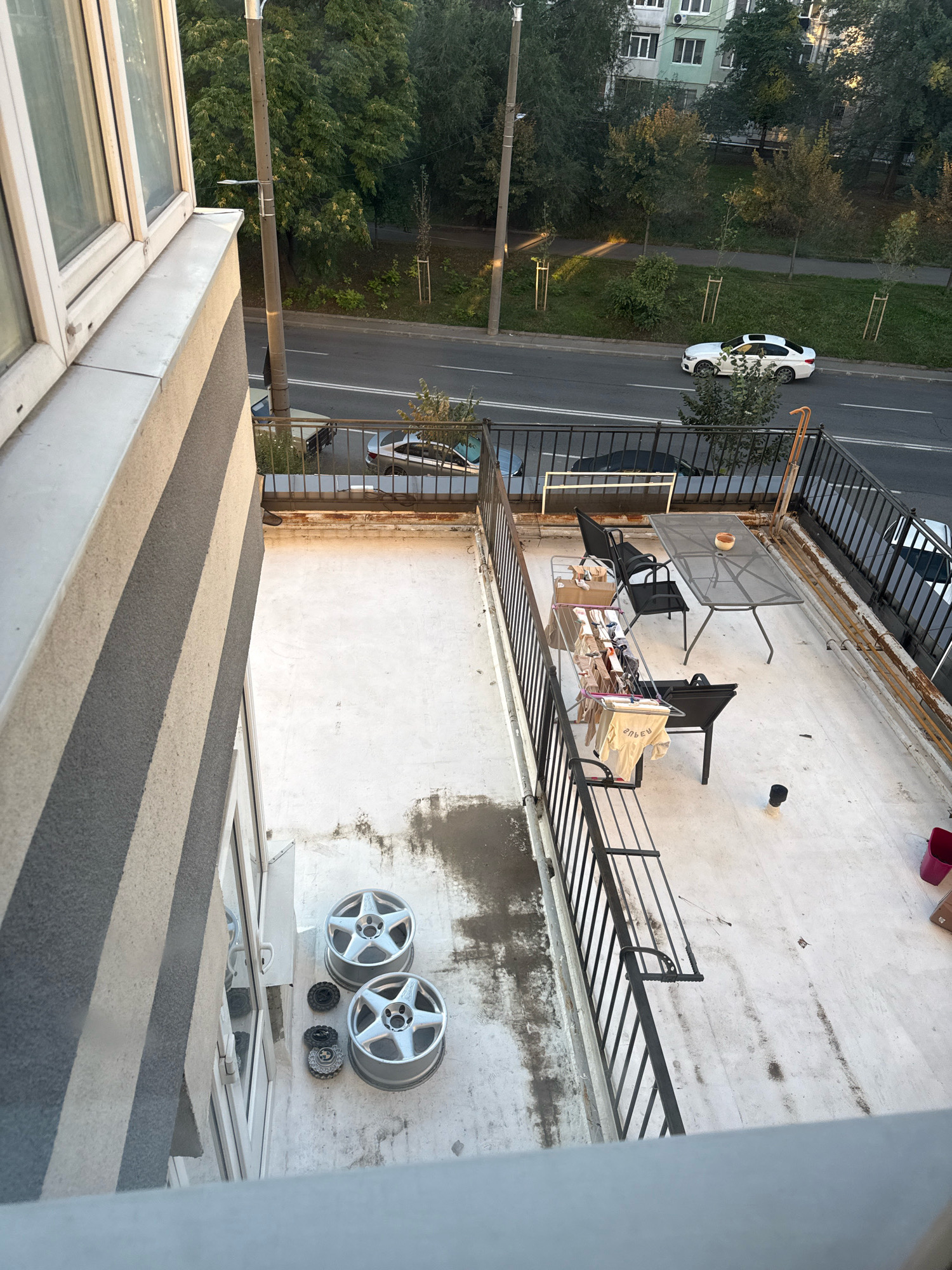 The image captures a view from a balcony looking down onto a paved balcony with various objects and a street with cars. The balcony is furnished with a metal table and chairs. A clothes drying rack with laundry is set up, and some cardboard boxes are present. Two car wheel rims are placed on the balcony floor. A small black pipe protrudes from the balcony floor, and a red bucket and a cardboard box are visible near the railing. The street below shows a white car driving, and other cars parked or moving. Trees line the street, and apartment buildings are visible in the background, suggesting an urban setting. The lighting indicates it is likely late afternoon or early morning. The overall scene appears to be a residential area in Chișinău, Moldova.
