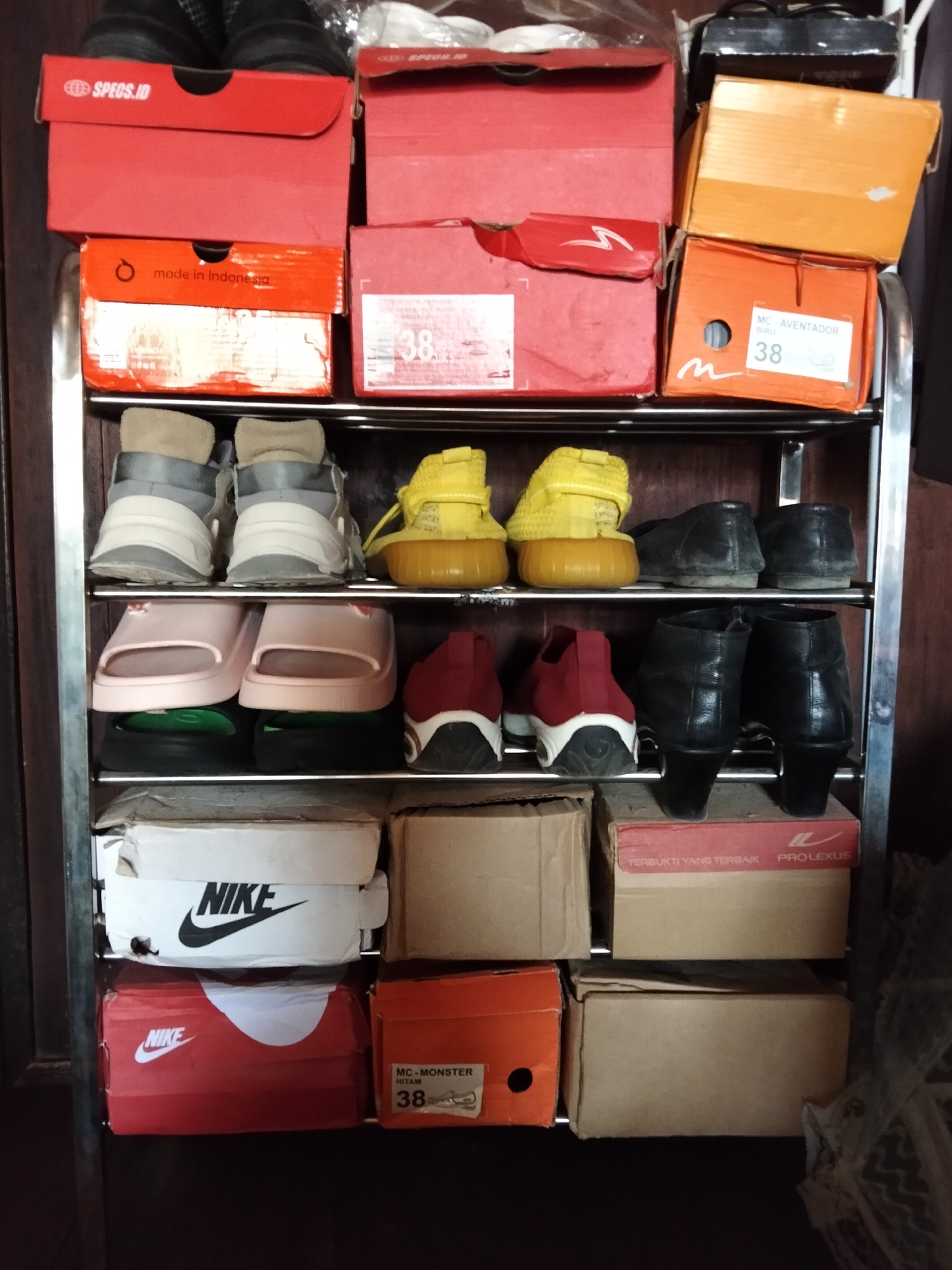 An indoor shot shows a metal shoe rack with several shelves filled with shoe boxes and shoes. The rack is located in Setia Jaya, Indonesia. The shoe boxes are of various colors and brands, including Nike, Specs, and Pro Lexus. Some shoes are visible, including sneakers, sandals, and boots. The background appears to be a wooden wall or cabinet.