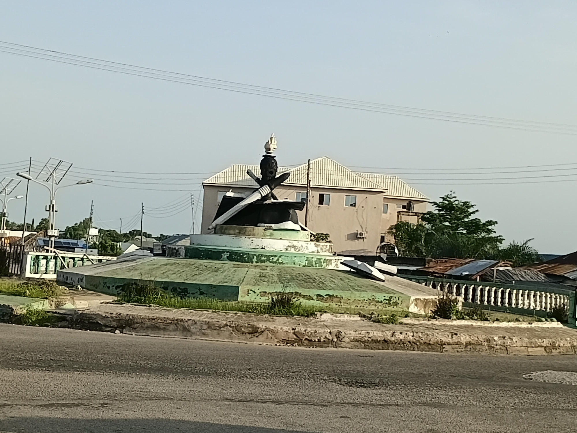 The image shows a monument at a roundabout in Jalingo, Nigeria. The monument is a statue on a tiered base. The statue is dark, possibly made of metal or stone, and appears to depict a figure wearing a crown and holding weapons, one of which is a large sword. The base of the monument is painted green and white, with some areas showing wear and peeling paint. Surrounding the monument is a paved area and some vegetation. In the background, there are buildings, including a multi-story building with a light-colored facade and a tiled roof, as well as trees and overhead power lines. The sky is clear and light blue. The overall scene suggests an outdoor public space in a city.