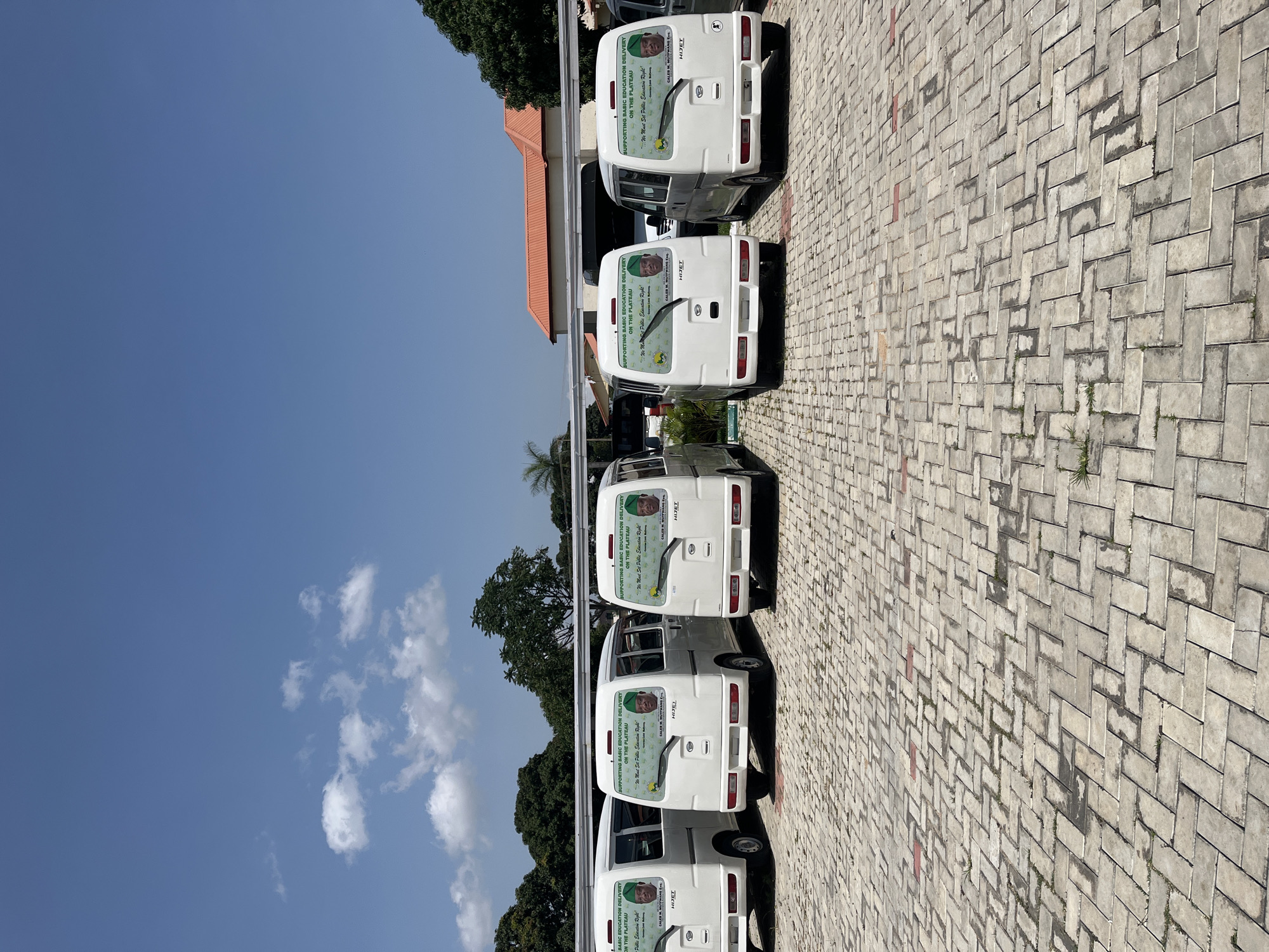 The image depicts a fleet of six white minivans parked on a paved lot in Jos, Nigeria. The vans are arranged in a line, facing away from the camera. Each van has a decal on its rear window that reads "SUPPORTING BASIC EDUCATION DELIVERY ON THE PLATEAU" and features two portraits of individuals, along with the text "We Must Set Public Education Right" and "CALEB M. MUTTWANG Esq.". The environment suggests an outdoor setting, possibly a parking area associated with a government building or educational institution, given the context of the decals. The sky is blue with scattered white clouds. The paving stones are arranged in a herringbone pattern. Lush green trees are visible in the background, and a building with an orange-tiled roof is also present. The overall scene is one of organization and a clear message of political or social support for education.