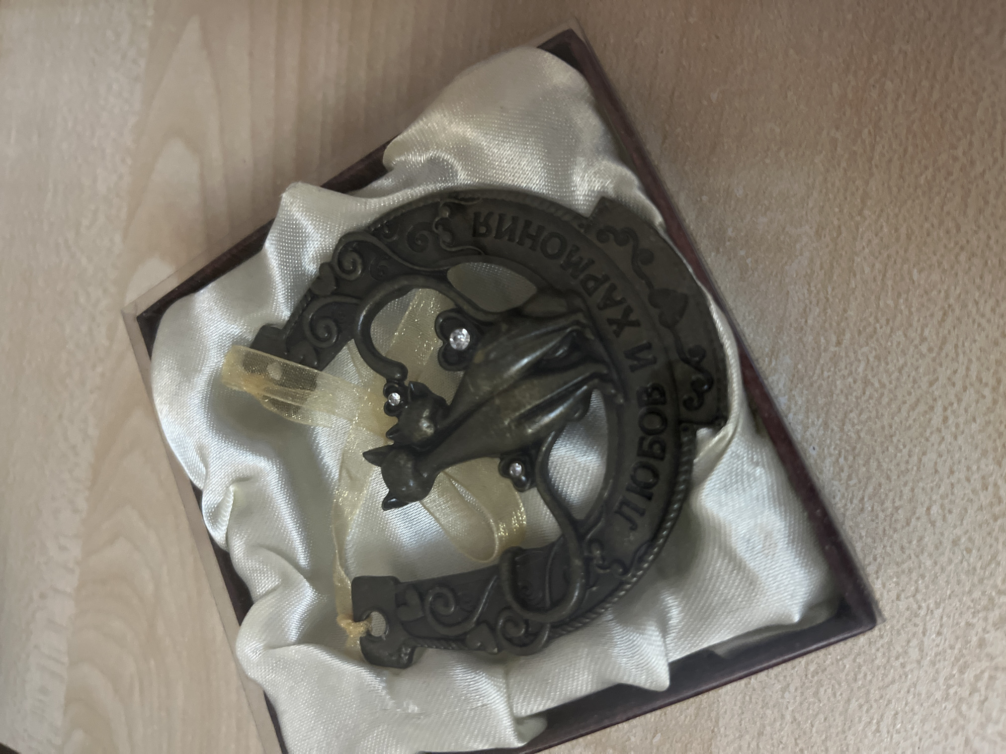 An antique horseshoe ornament with two cats and a ribbon is inside a dark brown box lined with white satin. The ornament is made of dark metal and has Cyrillic writing on it. The cats are facing each other, and the ribbon is gold. The box is on a wooden surface.