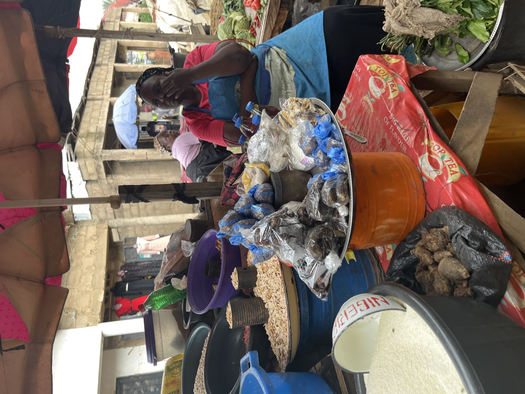 A street vendor sits under a pink and tan awning at a market stall in Jos, Nigeria. The vendor, a woman with dark skin, wears a pink t-shirt and a blue apron. She is looking down and appears to be rubbing her eye. In front of her are various goods for sale, including bags of dried goods, spices, and what appears to be rice or grain in a large black bowl. A red and white striped scoop is partially submerged in the white grains. A metal tray holds several small, individually wrapped packages of dark-colored items, possibly dried fish or meat. Behind her, other vendors and shoppers are visible, as well as stalls with clothing and produce. The overall scene depicts a vibrant, bustling market environment in an urban setting.