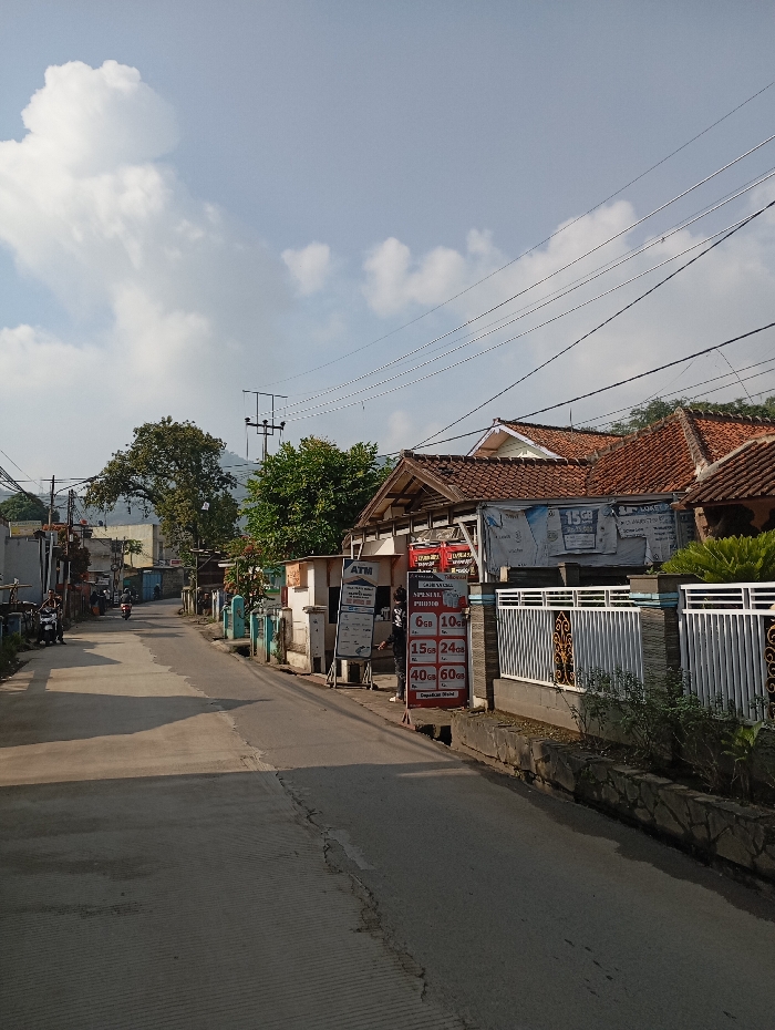 The image displays a daytime street scene in Situwangi, Indonesia. A two-lane asphalt road, partially shadowed, extends from the foreground into the background. On the right side of the road, a series of structures includes a building with a red tiled roof and another featuring an "ATM" sign. Adjacent to these buildings, multiple advertising boards are present; a prominent red vertical board lists data package prices, and a white board indicates "GADAI HP" (Pawn Phone) and "SPECIAL FOTO" services. A single individual dressed in dark clothing stands on the sidewalk next to the red advertising board. A white metal fence with decorative gold accents borders the property on the right, above a stone retaining wall. On the left side of the road, additional single-story buildings and parked motorcycles are visible. In the distant background, a person on a motorcycle moves away from the camera. The scene is traversed by numerous overhead power lines and utility poles. Large trees are interspersed along both sides of the road in the mid-ground, with distant, tree-covered hills forming the horizon. The sky is mostly clear with scattered white cumulus clouds.