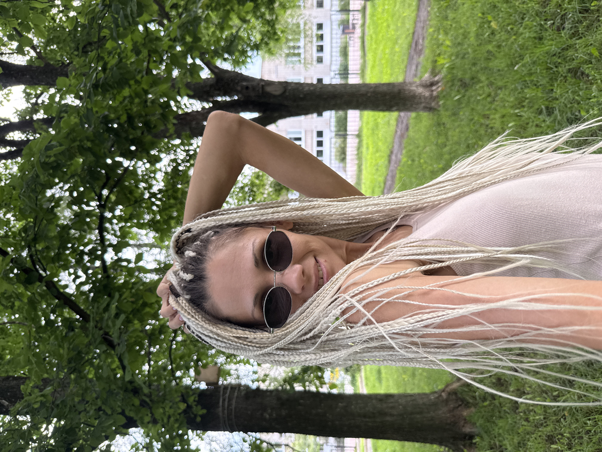 A white woman with blonde braids and sunglasses smiles while looking over her shoulder. She is standing outside in a grassy area with trees. A building and fence are visible in the background. The image was taken in Rusanovo, Russia.