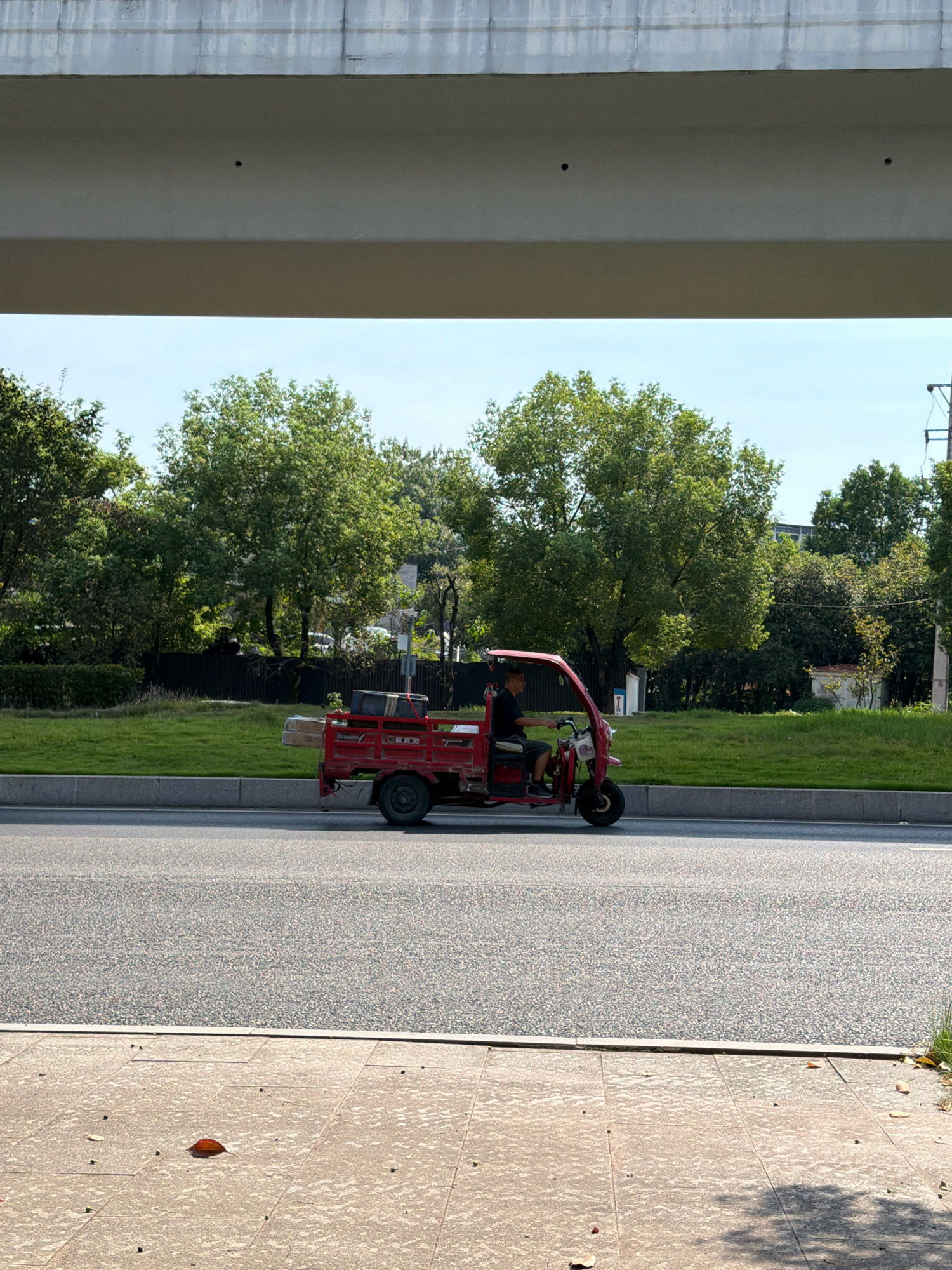 A man drives a red three-wheeled motorized vehicle down a paved road. The vehicle is loaded with several items, including what appears to be lumber and boxes. The man is wearing a dark t-shirt and shorts and is seated behind the steering wheel. Trees and grass line the road behind the vehicle. Overhead, a concrete overpass casts a shadow on the scene. The road is bordered by a sidewalk with scattered leaves. The sky is clear and blue. The scene is likely located in Jiangjin, China, as indicated by contextual information.