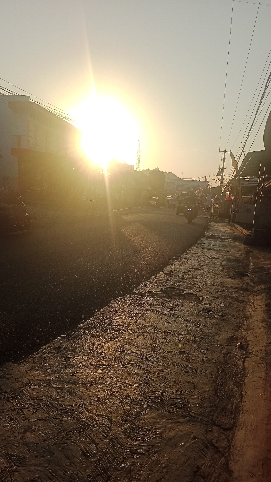 The image depicts a street scene in Karyamandala, Indonesia, during sunrise. The sun is prominently positioned in the upper left portion of the frame, casting a bright, warm glow that creates lens flares and long shadows across the asphalt road. The road is wet, reflecting the sunlight. On the left side of the road, a building with a white facade and an orange accent is visible, with a motorcycle parked in front. Further down the road, several vehicles, including motorcycles and cars, are present, suggesting active morning traffic. The right side of the road features a sidewalk or pavement with a textured, wavy pattern, and a row of buildings or stalls, some with awnings. Electrical wires are strung overhead. The sky is clear and pale yellow, typical of a sunrise. The overall atmosphere is one of a typical day beginning in a moderately busy urban environment.