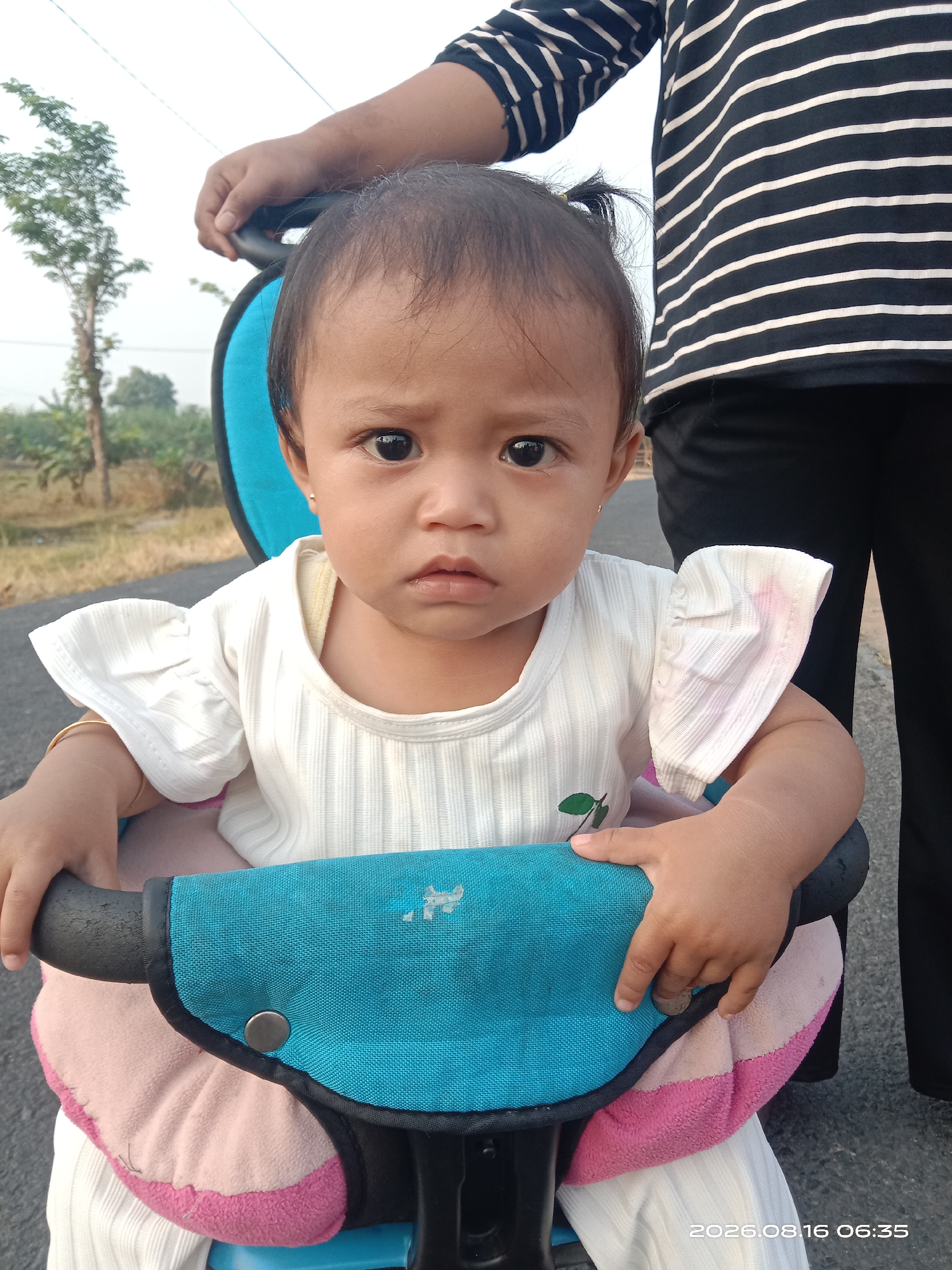 In Japanan, Indonesia, a young child sits in a stroller, looking directly at the camera with a serious expression. The child wears a white, ruffled shirt and has a small ponytail secured with an elastic band. A person's hand is visible in the upper left corner, holding the stroller's handle. The background shows a road with some trees and vegetation. The image also includes a timestamp: "2026.08.16 06:35".