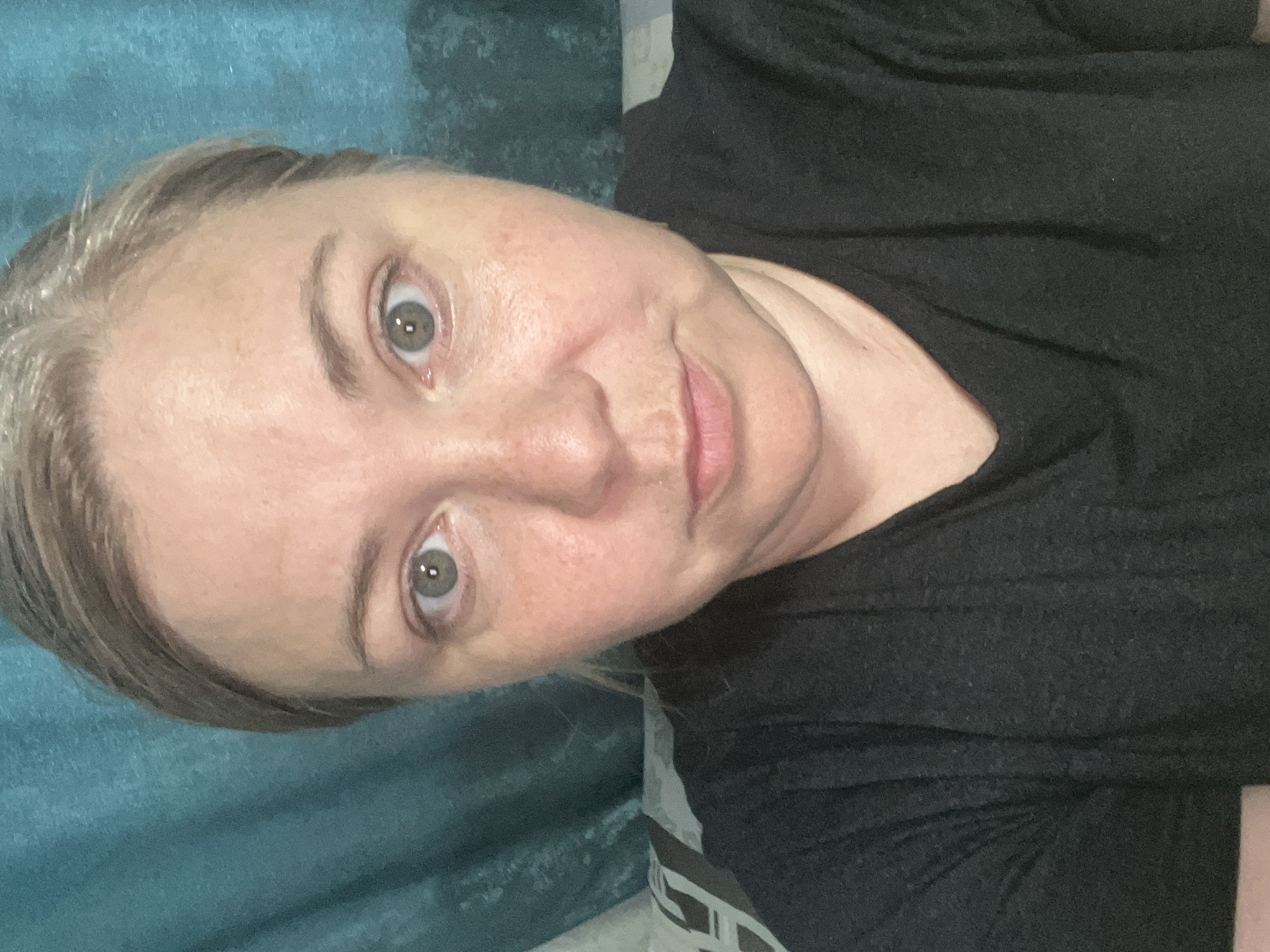 A close-up, head-and-shoulders selfie of a fair-skinned woman with light brown hair is captured against a teal-colored backdrop. Her gaze is directed towards the camera, and her expression is neutral. The background appears to be a curtain or drapery. The image was taken in Foki, Russia.