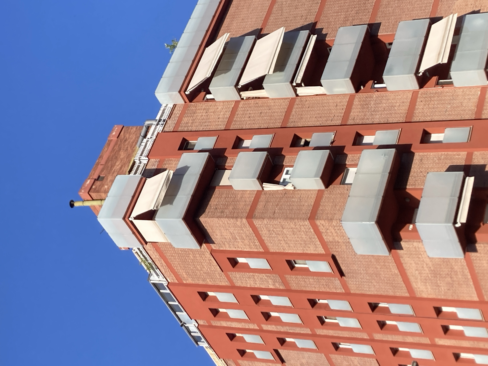 This is a low-angle, outdoor photograph of a modern building with a brick facade, seen against a clear blue sky. The building's design features repeating rectangular window openings with lighter-colored frames, interspersed with darker brickwork. Above some of these windows are protruding, box-like structures with translucent panels, possibly awnings or sunshades. The building has a contemporary architectural style, with clean lines and geometric shapes. There are also some retractable awnings extended over some of the windows. The lighting suggests it is daytime, likely mid-morning or afternoon, given the strong shadows. The clear blue sky indicates fair weather. The perspective of the photograph, looking upwards, emphasizes the height and scale of the building. No people are visible in the image. There is no visible text. The scene is consistent with an urban setting, possibly a residential or commercial building in a city like Barcelona, Spain, known for its modern architecture.