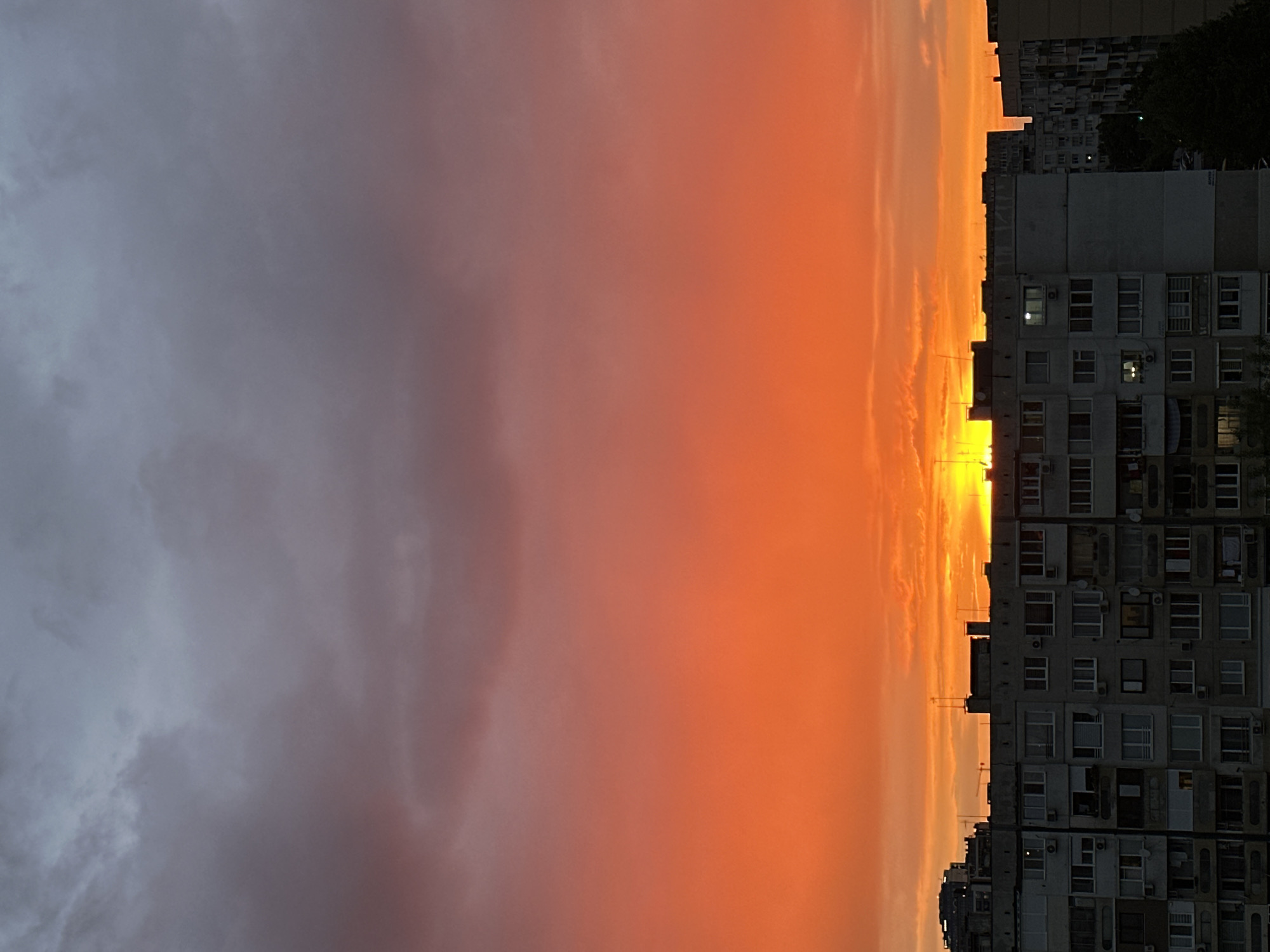 The image shows an orange sunset sky over the city of Sofia, Bulgaria. A tall apartment building with many windows dominates the right side of the frame. The building is likely located on the outskirts of the city, as evidenced by the open sky. The sky is predominantly orange near the horizon, transitioning to gray clouds on the left. The sun is obscured behind the apartment buildings, but its bright light creates a silhouette effect. There are no people visible in the frame.