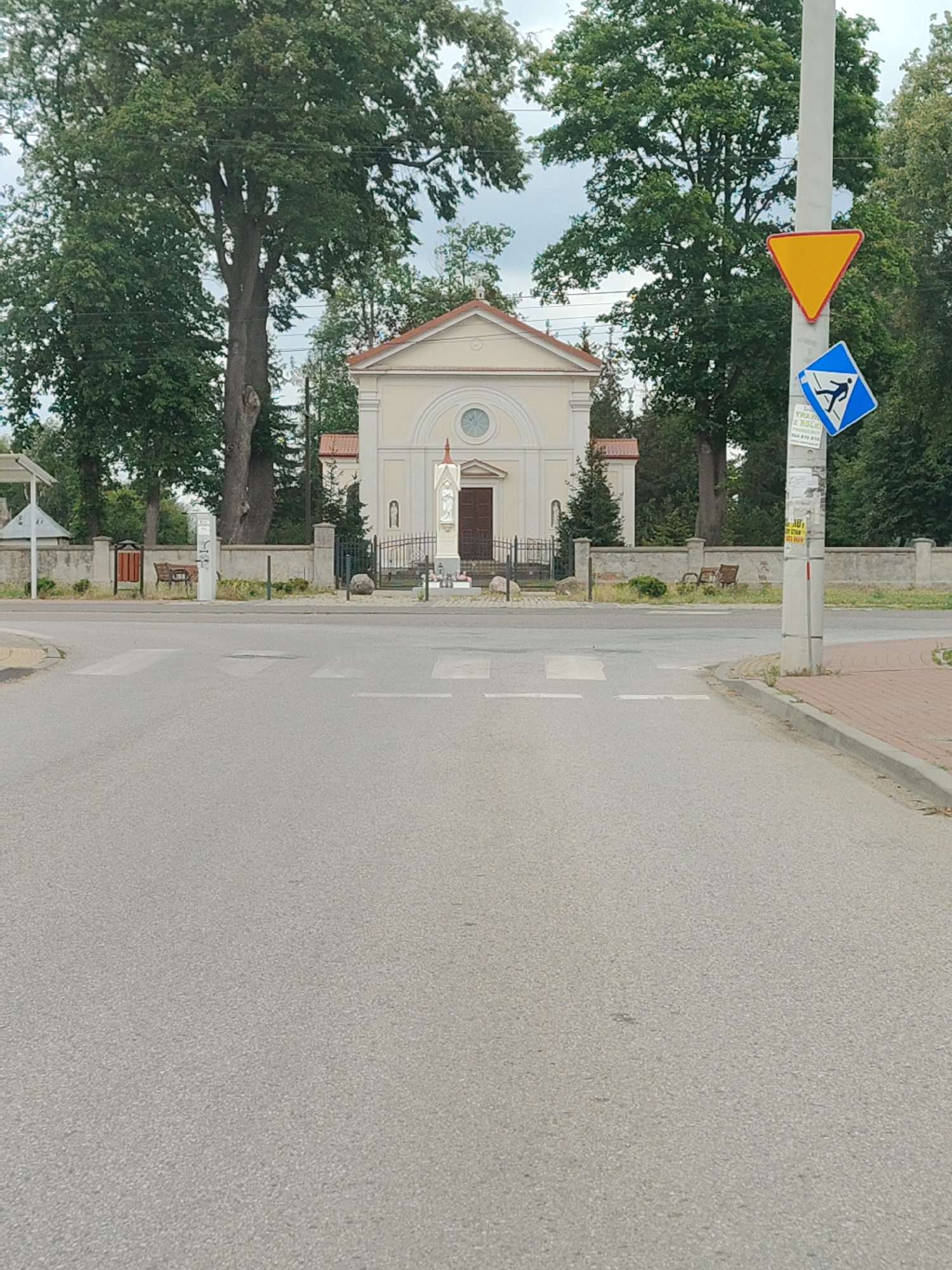 The image shows a street scene in Łomna, Poland. In the background, a small, cream-colored building with a red-tiled roof stands behind a low stone wall. The building has a central arched entrance and a circular window above it. A statue of a robed figure stands to the right of the entrance. Two large trees with dense green foliage frame the building on either side.
In the foreground, a paved road with a pedestrian crossing is visible. A light-colored utility pole is situated on the right side of the road, displaying two traffic signs: a yellow triangle pointing downwards and a blue square with a white silhouette of a person walking. The area behind the wall appears to be a grassy verge with some bushes and two wooden benches. The sky above is overcast.