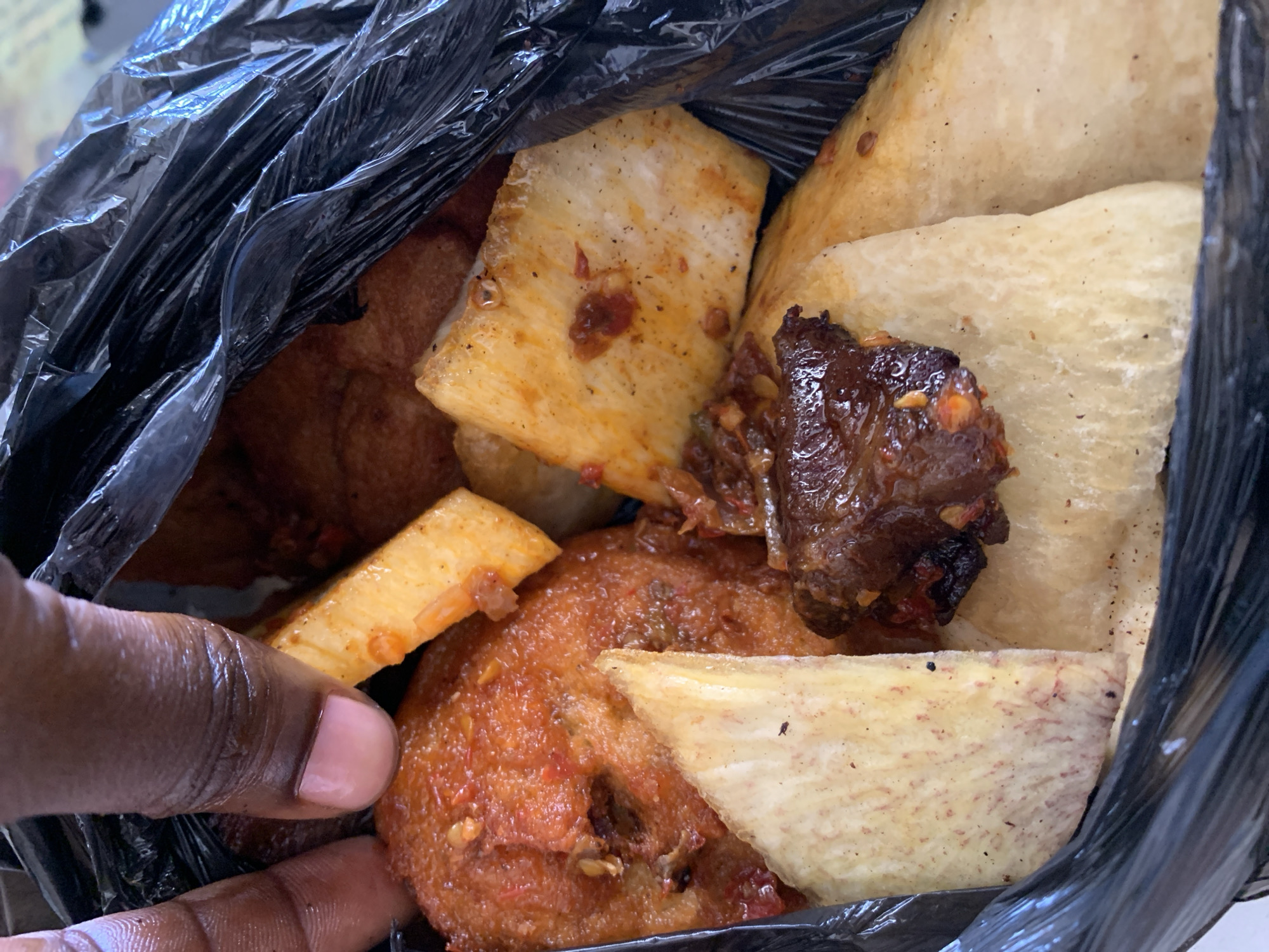 A close-up shot inside a black plastic bag reveals a collection of assorted African street food. The food includes fried yam pieces of varying sizes and colors from light tan to golden brown with a spicy coating, and dark pieces of cooked meat, possibly beef. A portion of a brown-skinned hand with a manicured nail is visible at the lower left edge of the bag. There is no information to indicate that this image was taken in the city of Kabong, Nigeria.
