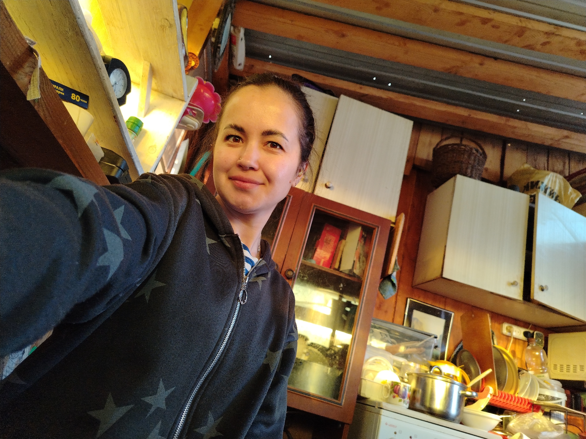 A selfie photograph shows a woman in an interior cluttered with household items. The woman is facing the camera with a slight smile. She has brown hair pulled back, and is wearing a black zip-up hoodie with gray stars. A silver nose ring is visible.
Behind her is a crowded kitchen or storage area. There is a wooden cabinet with a glass door, white cabinets, a microwave, pots, pans, dishes, and various other kitchen utensils and supplies. A clock is visible on a shelf on the upper left. The ceiling appears to be made of wood planks.
There is no information in the image to pinpoint a location within Noviy, Russia.