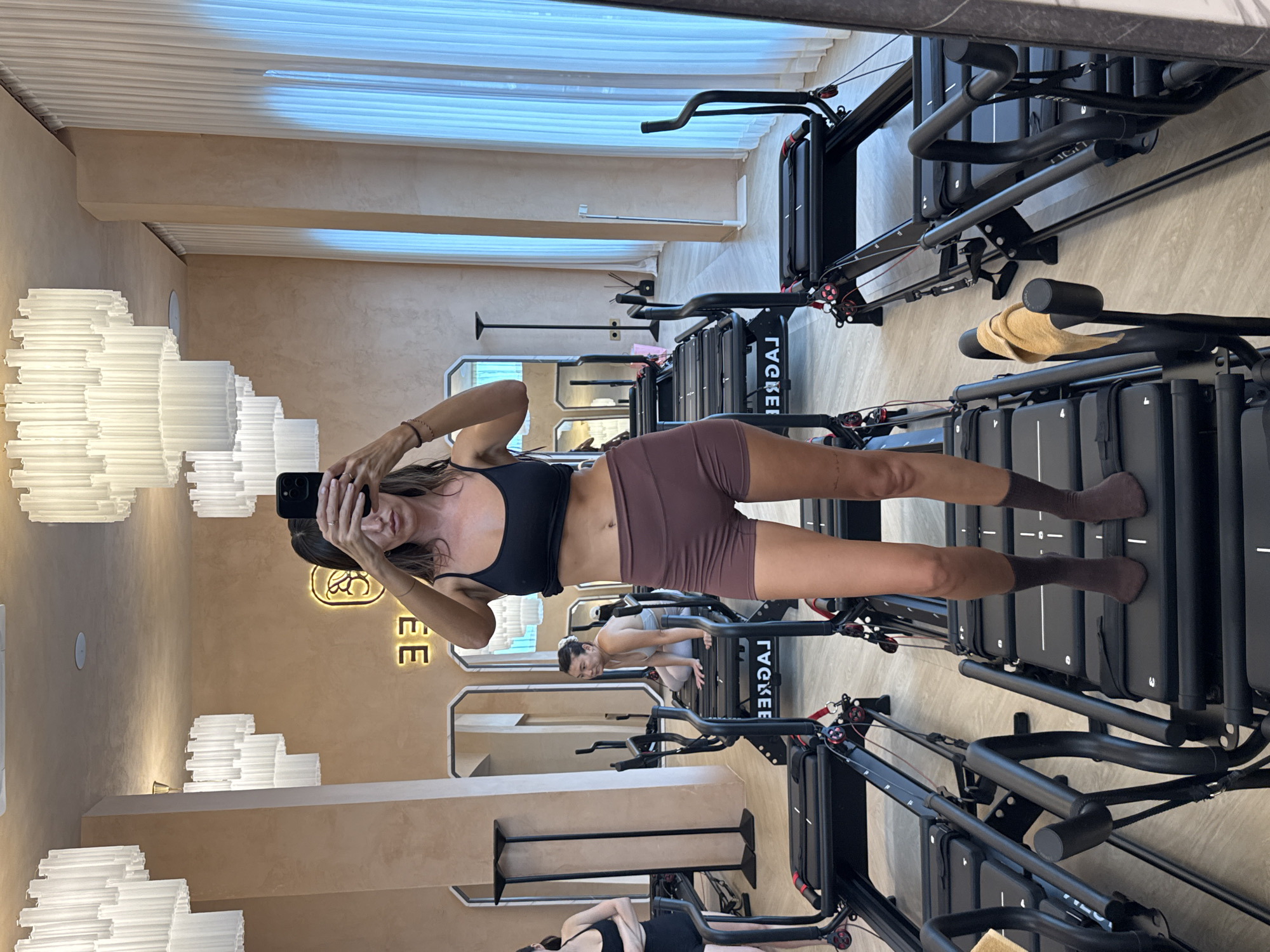 The image captures a mirror selfie taken by a woman within a modern fitness studio, likely a Pilates or Lagree Fitness establishment. The scene, which appears rotated 90 degrees counter-clockwise, shows the woman reflected in a large mirror, holding a dark-colored smartphone to take the picture, with her face partially obscured. She has long, dark hair and is dressed in a black sports bra, brown/mauve athletic shorts, and dark socks. A ring is visible on her left ring finger, and a dark bracelet adorns her left wrist.
The studio features several black, contemporary fitness machines, resembling Pilates reformers or Lagree Megaformers, arranged in rows. The brand name "LACKEE" is visible (reversed) on the side of some machines. A small, light-colored towel is draped over a handle on one of the machines in the foreground. In reflections from other mirrors in the background, at least two other individuals are visible. One woman, in light-colored activewear, is seen on a machine, seemingly engaged in or preparing for a workout.
The interior boasts warm, light brown or beige textured walls, complemented by distinctive white, tiered, sculptural light fixtures mounted on the walls and ceiling. The floor is light-colored. A neon sign, partially visible on a wall, spells "COFFEE" (reversed) with a circular logo above it, suggesting the studio might also house a cafe. The overall setting is clean and well-lit, indicating a professional and stylish fitness environment in North Kuta, Indonesia. The time of day is not discernible from the indoor lighting.