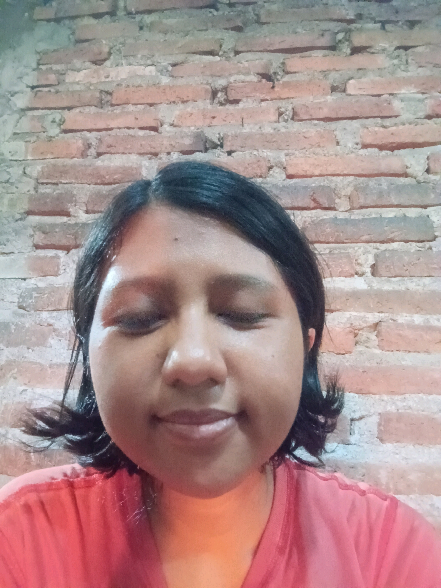 A young woman with dark hair and brown eyes is pictured from the chest up, facing forward with her eyes closed. She is wearing a salmon-colored t-shirt. Her skin appears slightly flushed, and her hair is dark and shoulder-length, with some strands falling forward around her face. The background is a brick wall, with visible mortar between the bricks. The lighting in the image is warm, casting a soft glow on the woman's face and shoulders. The overall impression is a casual, candid portrait. The location is specified as Canga'an, Indonesia.