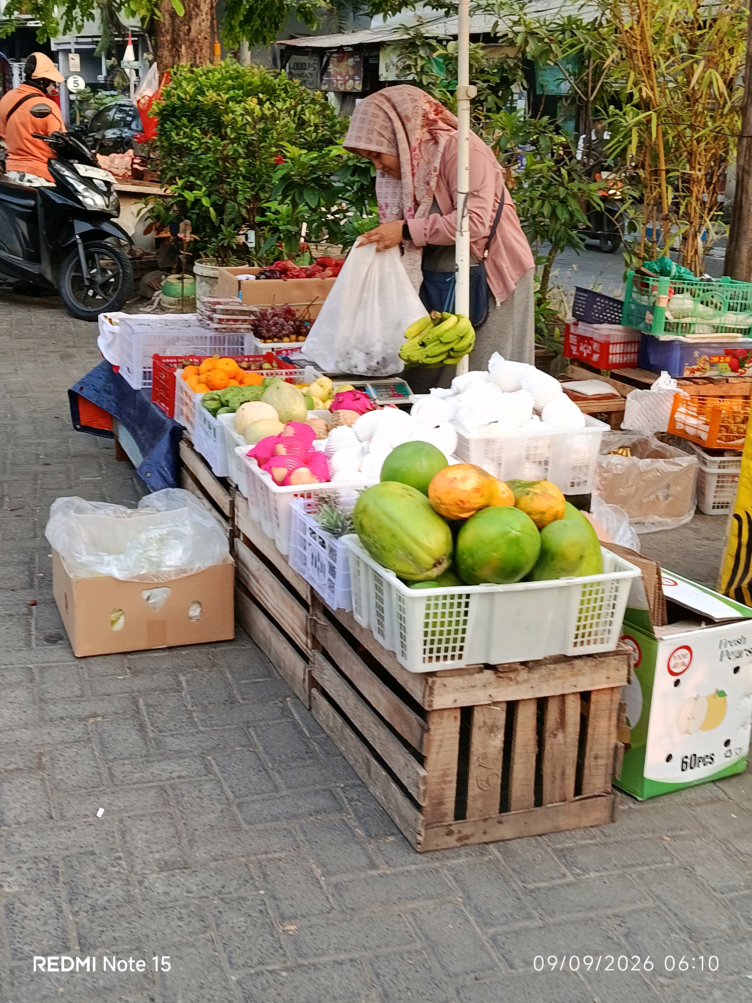 A street market scene in Keboansikep, Indonesia features a woman in a light pink hijab and jacket standing behind a display of fresh fruits. She is holding a white plastic bag, appearing to be in the process of bagging items. The fruits are arranged in white plastic crates, with visible papayas, bananas, oranges, and what appear to be melons and pomegranates. Some of the fruit is also displayed in wooden crates and brown cardboard boxes. In the background, a motorcycle is parked, and a person in an orange shirt and cap is seated on it. Lush green foliage and bamboo stalks are visible behind the market stall. The ground is paved with grey interlocking bricks. A portion of a box with the text "Fresh Pears" is visible on the right side of the frame. The timestamp "09/09/2026 06:10" and "REDMI Note 15" are overlaid on the bottom of the image.