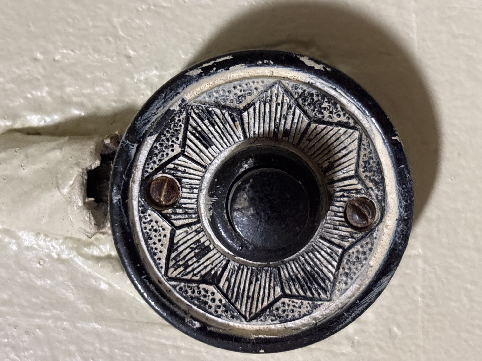 The image shows a close-up, eye-level view of an old, ornate, black and white doorbell button mounted on a textured, cream-colored wall. The doorbell has a circular, dark metal outer rim, and a decorative, lighter-colored circular faceplate with a starburst pattern. The starburst pattern features alternating sections of fine dots and radiating lines. In the center of the faceplate is a black, circular button, recessed slightly. Two brown, Phillips-head screws are visible, securing the doorbell to the wall, one at the top and one at the bottom of the faceplate. Above the doorbell, a section of white, tape-like material is adhered to the wall, appearing worn and slightly torn, with some evidence of old wiring or insulation beneath it. The wall itself has a rough, stucco-like texture. The lighting casts a soft shadow to the right of the doorbell.
The specific location of this doorbell within the city of Tallinn, Estonia, cannot be determined from the provided image alone.