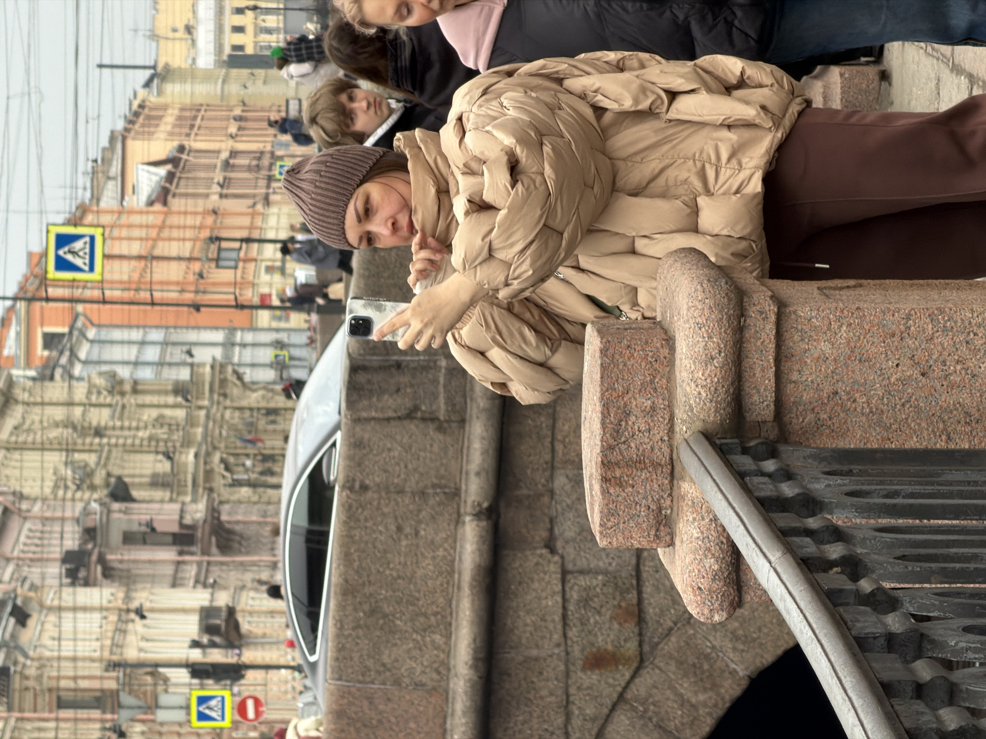 An urban street scene in Saint Petersburg, Russia, depicts a woman photographing on a bridge over the Griboyedov Canal. The primary subject, a woman positioned mid-frame, wears a brown knit beanie, a voluminous beige puffer jacket, and dark brown trousers. She holds a silver smartphone horizontally with both hands, pointed towards her left. Her expression is focused. To her right and slightly behind, a young man with light brown hair looks towards the camera. The immediate foreground features a brown-granite bridge parapet and a dark, ornate metal railing. A modern, dark-colored car is partially visible on the street behind the granite embankment. A traffic pole displaying a blue pedestrian crossing sign and a red circular regulatory sign is visible above the car. The background consists of characteristic multi-story Saint Petersburg buildings in various pastel shades (beige, orange, yellow), featuring ornate architectural details, under an overcast sky. Several blurred pedestrians are visible in the distant background.