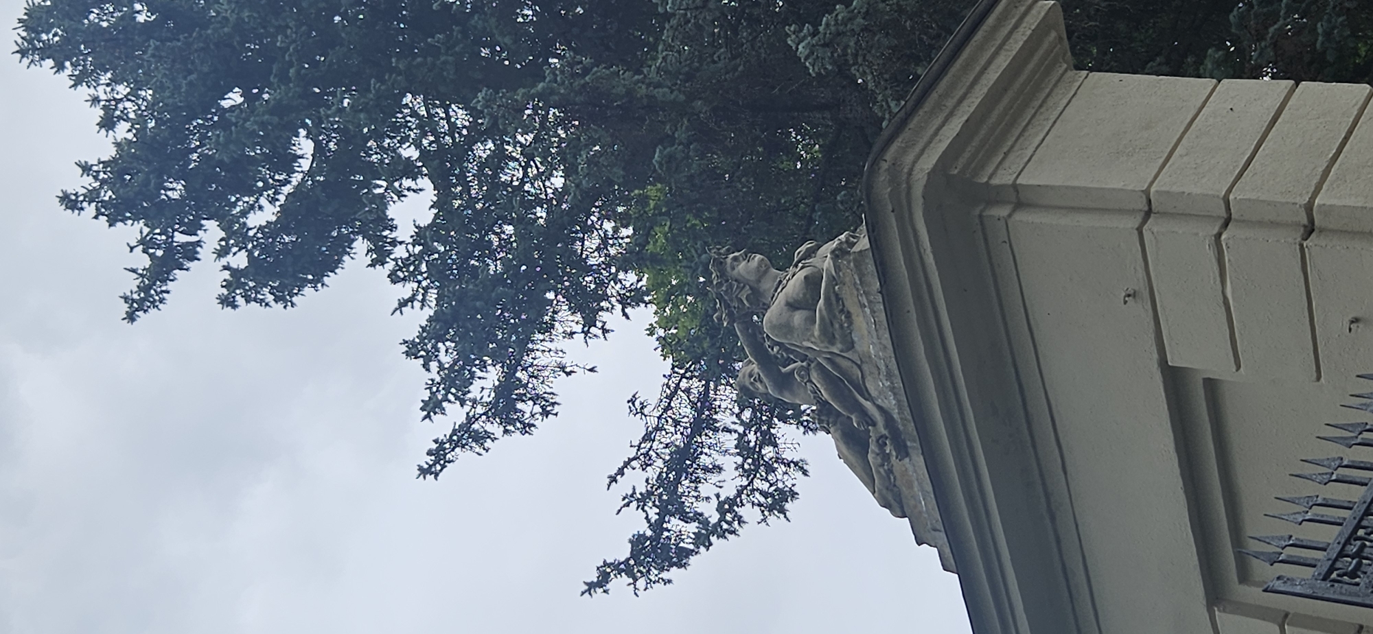 This image depicts a stone sculpture situated atop a light-colored pillar or wall. The sculpture features two figures: a larger, draped female figure with a crowned head, and a smaller child-like figure seated behind and above her, with an arm extended. Both figures appear to be lying down. Behind the sculpture, a large evergreen tree dominates the frame, with its branches reaching towards a cloudy sky. The pillar itself has decorative moldings. In the foreground, part of a dark metal fence is visible. This scene is located at the entrance of the historic Grodno State Museum of History and Religion in Hrodna, Belarus.
