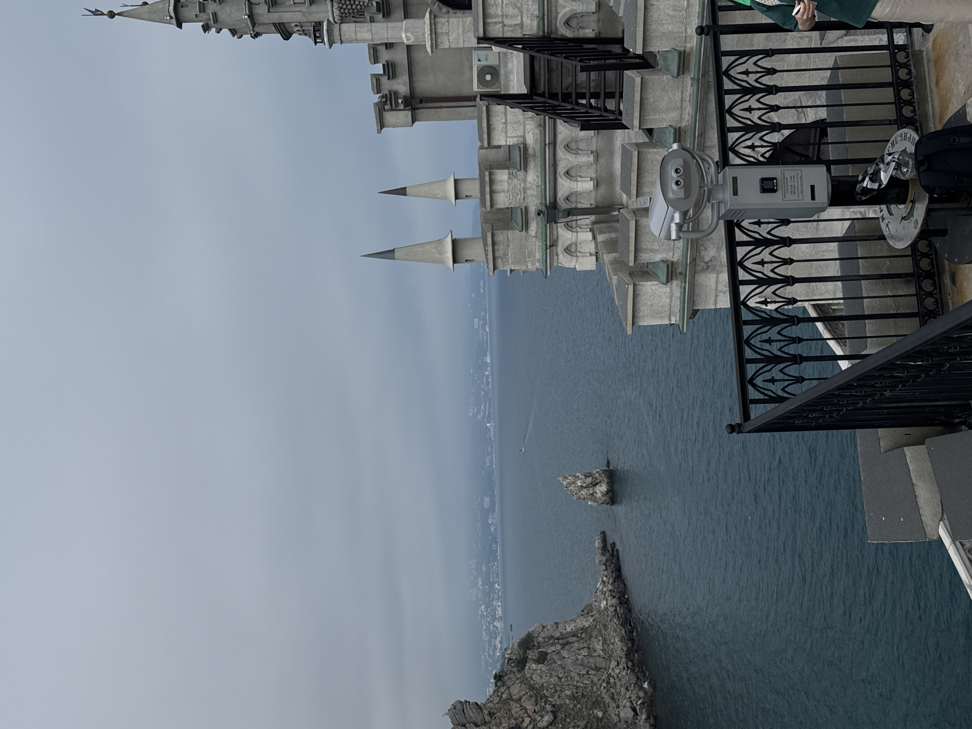 This is an outdoor shot of the Swallow's Nest castle in the Гаспринский поселковый совет area of Ukraine, taken from an elevated perspective. The castle's exterior, crafted from light-colored stone, is prominently visible, featuring multiple turrets and decorative elements, indicative of its architectural style. The background showcases the dark blue sea meeting the horizon under a muted gray sky. There is also rocky formations below the castle and in the distance. A stationary binocular stands on the castle's edge, adjacent to a black, decorative railing. A person wearing a green jacket is partially visible on the right side of the frame.
