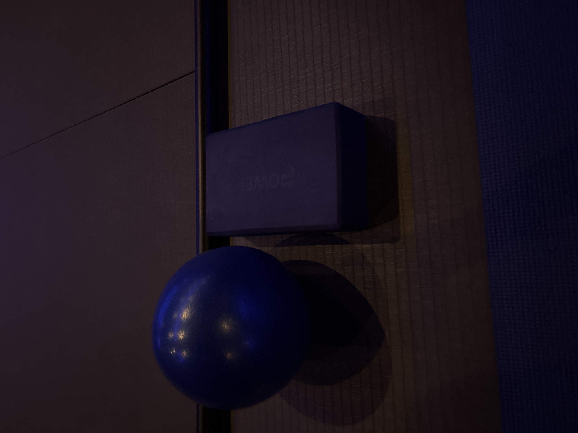 A close-up shot depicts a blue yoga block with the word "POWER" visible on its side, placed above a blue exercise ball. They are positioned against a textured wall. The scene appears to be indoors.