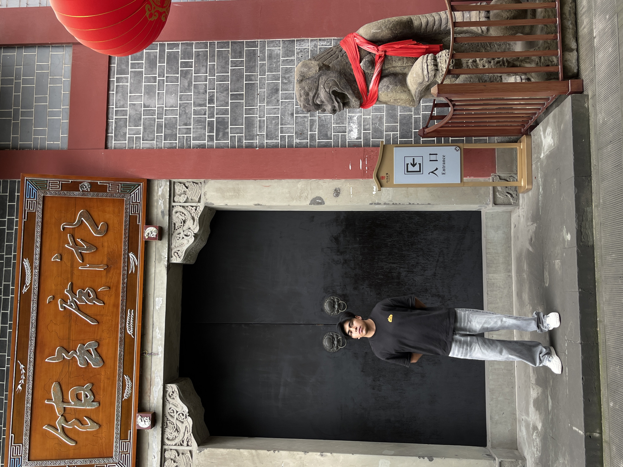 A young man in a black t-shirt, light grey jeans, and white sneakers poses in front of a large, closed, black wooden door with ornate metal handles. The location is in the Yuzhong District of China. The door is flanked by traditional Chinese architectural elements, including ornate stone carvings and a wooden plaque with Chinese characters. Above the door, there's a stone sculpture of a mythical creature with a red cloth tied around its neck and a sign that reads "Entrance" in both English and Chinese characters. A red lantern is hung above the entrance. The background features brickwork in a grey and white pattern. The image is oriented vertically.