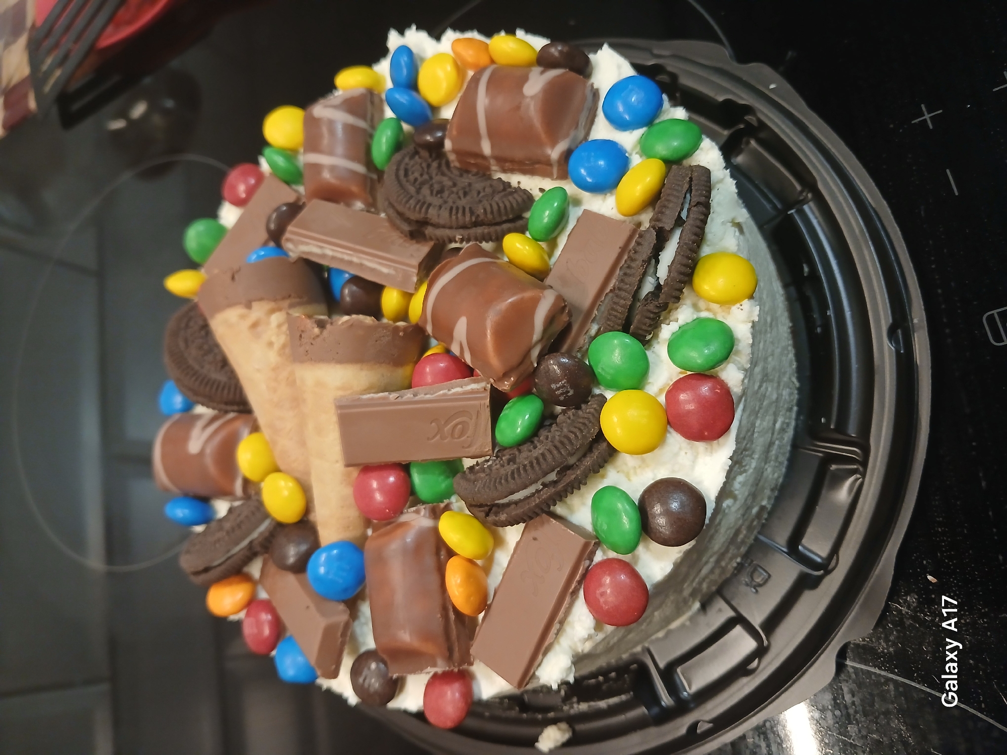 An overhead shot captures a lavishly decorated round dessert, likely a cake, presented in a black plastic container. The cake is covered in a substantial layer of white cream or frosting and is densely topped with a variety of confectionery items. This scene is set on a dark, reflective surface, possibly a kitchen countertop or stovetop, in Yekaterinburg, Russia.
The vibrant decorations include numerous candy-coated chocolates in a spectrum of colors: red, yellow, green, blue, and orange. Several types of chocolate bars are arranged on the cake; some are plain milk chocolate, while others feature a distinct white stripe, similar to Kinder chocolates. One visible chocolate bar clearly displays the text "Fox". Chocolate sandwich cookies, resembling Oreos, are also integrated into the design. Two waffle cones, typically used for ice cream, are placed upright on the cake, contributing to its multi-textured appearance. Small, round brown chocolate candies are scattered among the other toppings.
The black plastic container has a ridged perimeter, and one of the detailed crops reveals a recycling symbol, specifically for plastic, embossed on its side. The dark background surface features faint white markings, possibly indicators on a stovetop. The text "Galaxy A17" is visible as a watermark in the bottom right corner of the original image, indicating the camera used. The image solely focuses on the dessert; no people are visible, and no specific event is directly depicted, though the elaborate decoration suggests a celebratory occasion or a special treat.