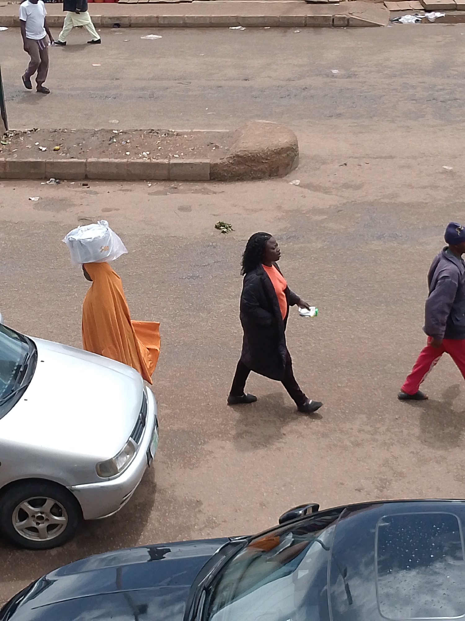 This is an eye-level, full-shot, outdoor, daytime, high-angle photograph of a street scene in Bukuru, Nigeria. In the foreground, the black roof and windshield of a car are visible. To the left, the silver hood and front wheel of another car are in focus. In the middle ground, three individuals are walking on the paved road. The leftmost individual is wearing an orange robe and carrying a white object on their head, wrapped in plastic. The middle individual is wearing a black coat over an orange shirt and dark pants, and is holding a small white object in their right hand. The rightmost individual is wearing a blue hat, a gray jacket, and red pants with a white stripe. The background consists of a paved road with some debris and a sidewalk with a curb.