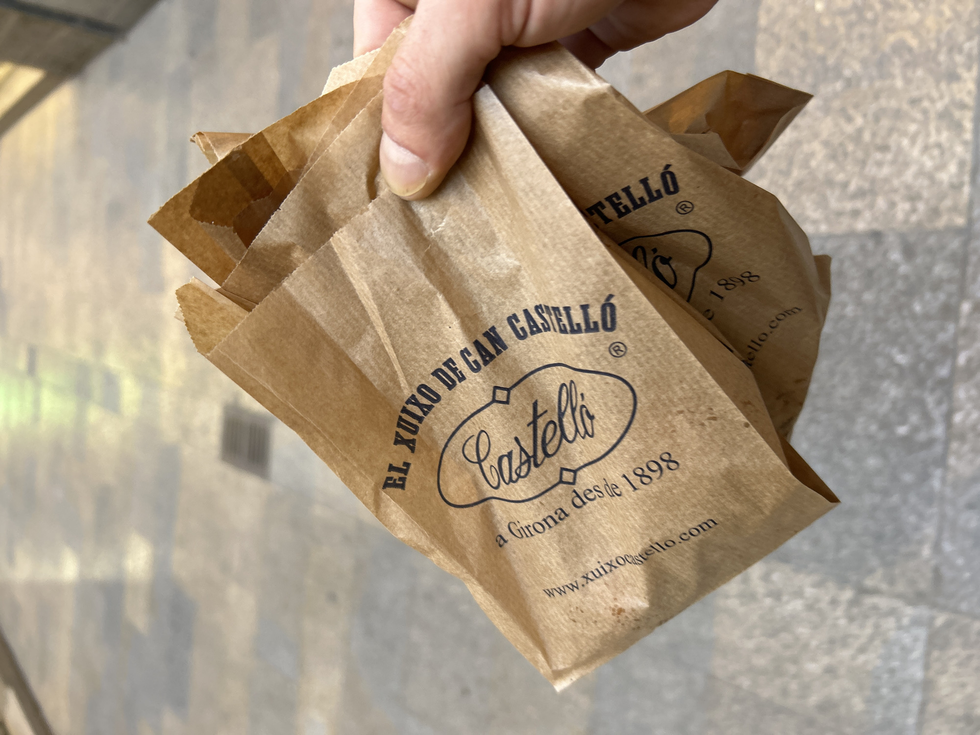 A close-up, eye-level, outdoor shot shows a hand holding a stack of crumpled brown paper bags in Girona, Spain. The bags have black text and a logo that reads "Castelló" within an oval outline, along with the text "EL XUIXO DE CAN CASTELLÓ" and "a Girona desde 1898." The web address "www.xuixocastello.com" is also visible. The background consists of a light-colored, tiled or paved pedestrian walkway with some visible drain covers. The lighting suggests it is daytime.