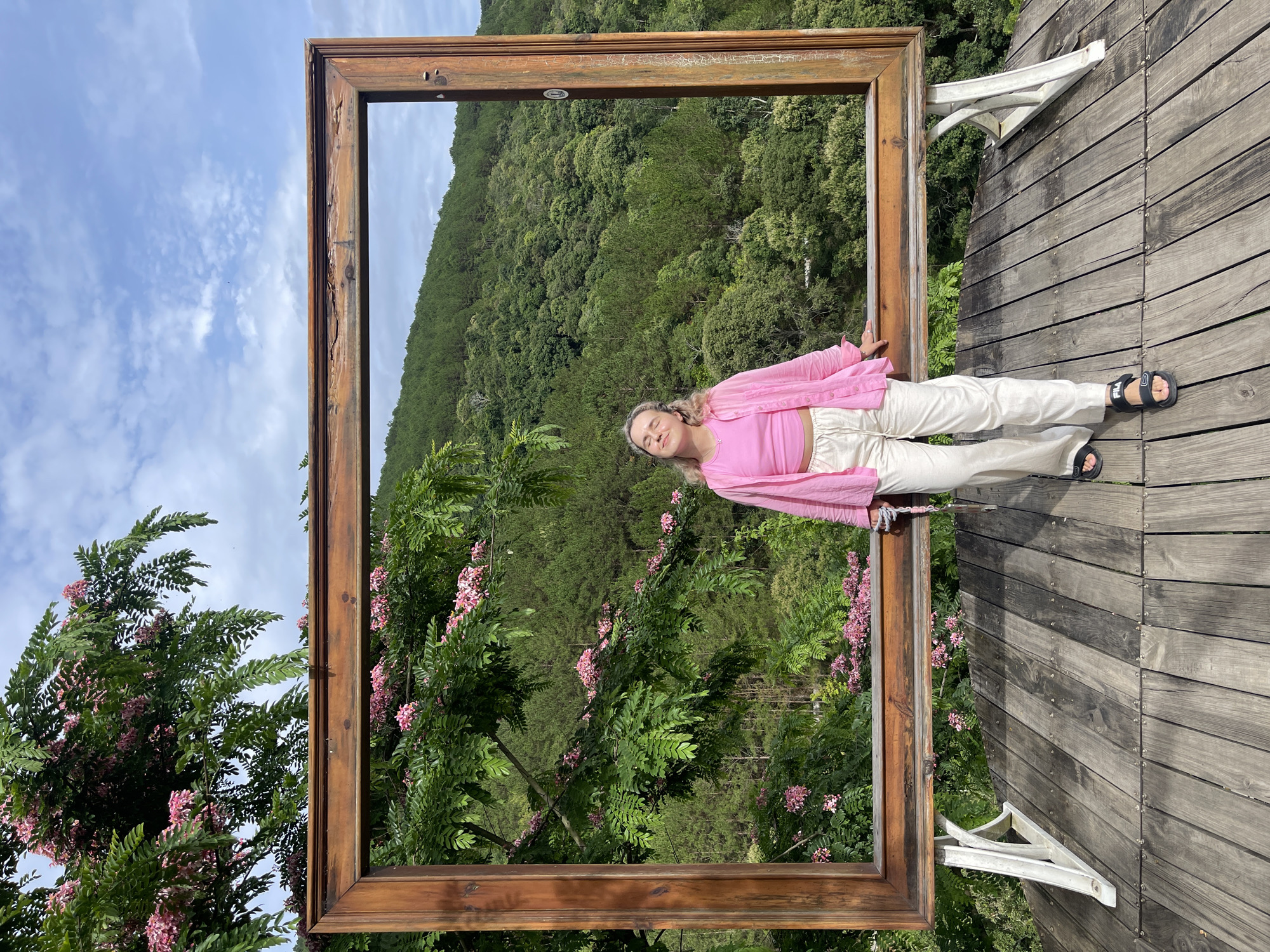 The image shows a woman in a pink top and cream pants lying on a wooden deck within a large, wooden frame. The frame is positioned to capture a view of a forested hillside, a glimpse of a cloudy sky, and flowering trees. The deck is weathered wood, secured with visible screws. The frame itself has rustic detailing. The photo appears to have been taken in daylight in Thôn 5, Vietnam, as a location tag implies. The angle of the photo shows it taken by someone looking down on the woman.