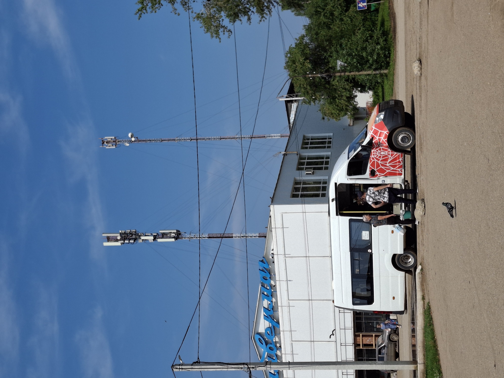 In Elovo, Russia, a white minibus with a red abstract design on its front quarter is parked on a paved road. Two individuals stand near the open passenger door of the minibus, one holding a bucket. A woman in a blue dress walks away from the vehicle towards a car. In the background, a white building with blue lettering adorns its facade, and two tall telecommunications towers rise against a clear blue sky. The environment appears to be an outdoor public space, possibly near a commercial or residential area, with trees and power lines visible. A pigeon is present on the road in the foreground.