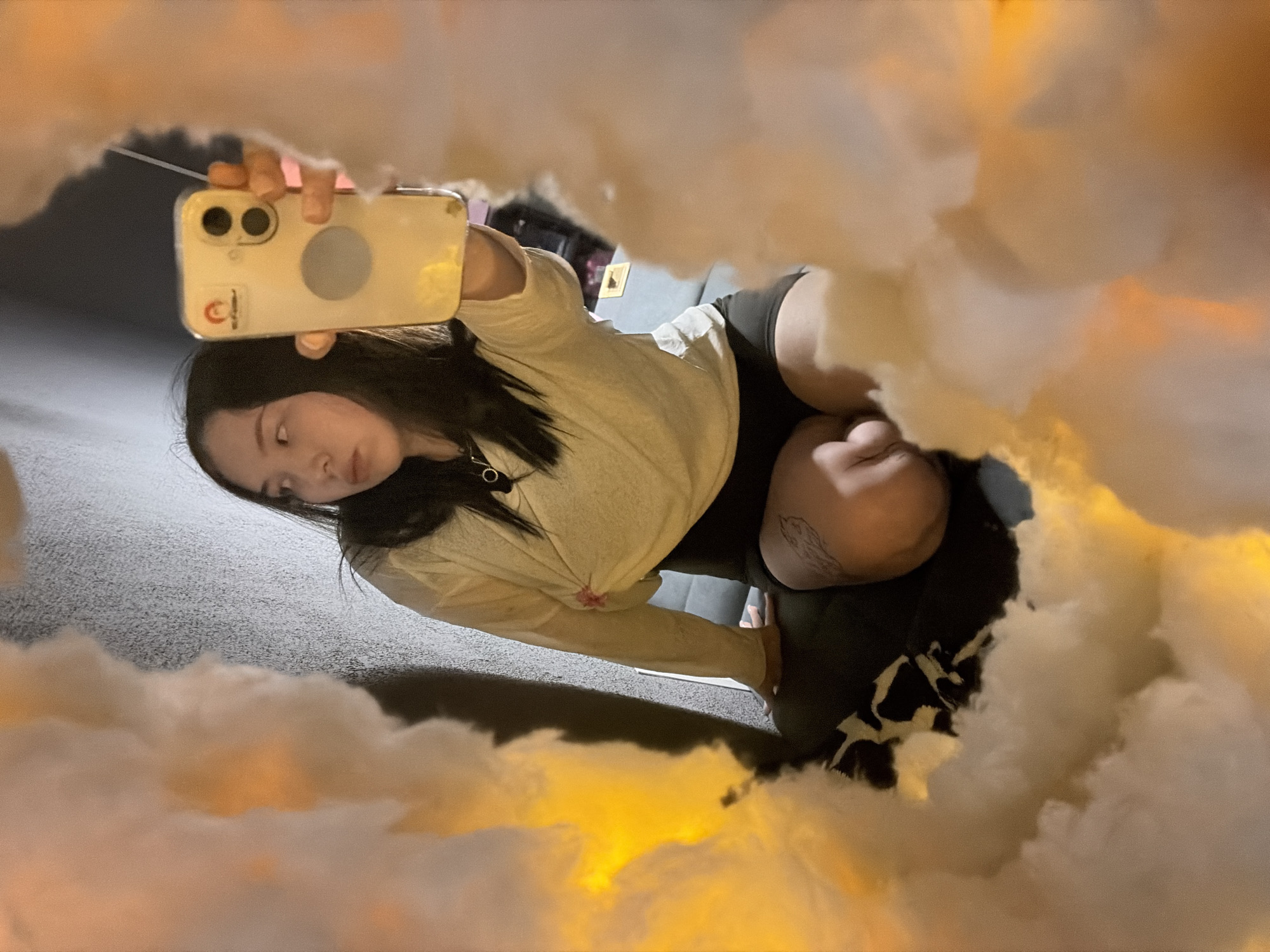 A young Asian woman is reflected in a mirror, taking a selfie with her smartphone. She is sitting down, wearing a light-colored long-sleeved shirt and dark shorts. There is a tattoo on her right thigh. The background is dark and textured, with some blurred objects visible. The foreground of the mirror is partially obscured by fluffy, cloud-like decorations that are lit with warm, orange light, creating a dreamy or ethereal effect. The scene is likely indoors. The image does not provide any specific information or visual cues to indicate a location in Chaikovsky, Russia.