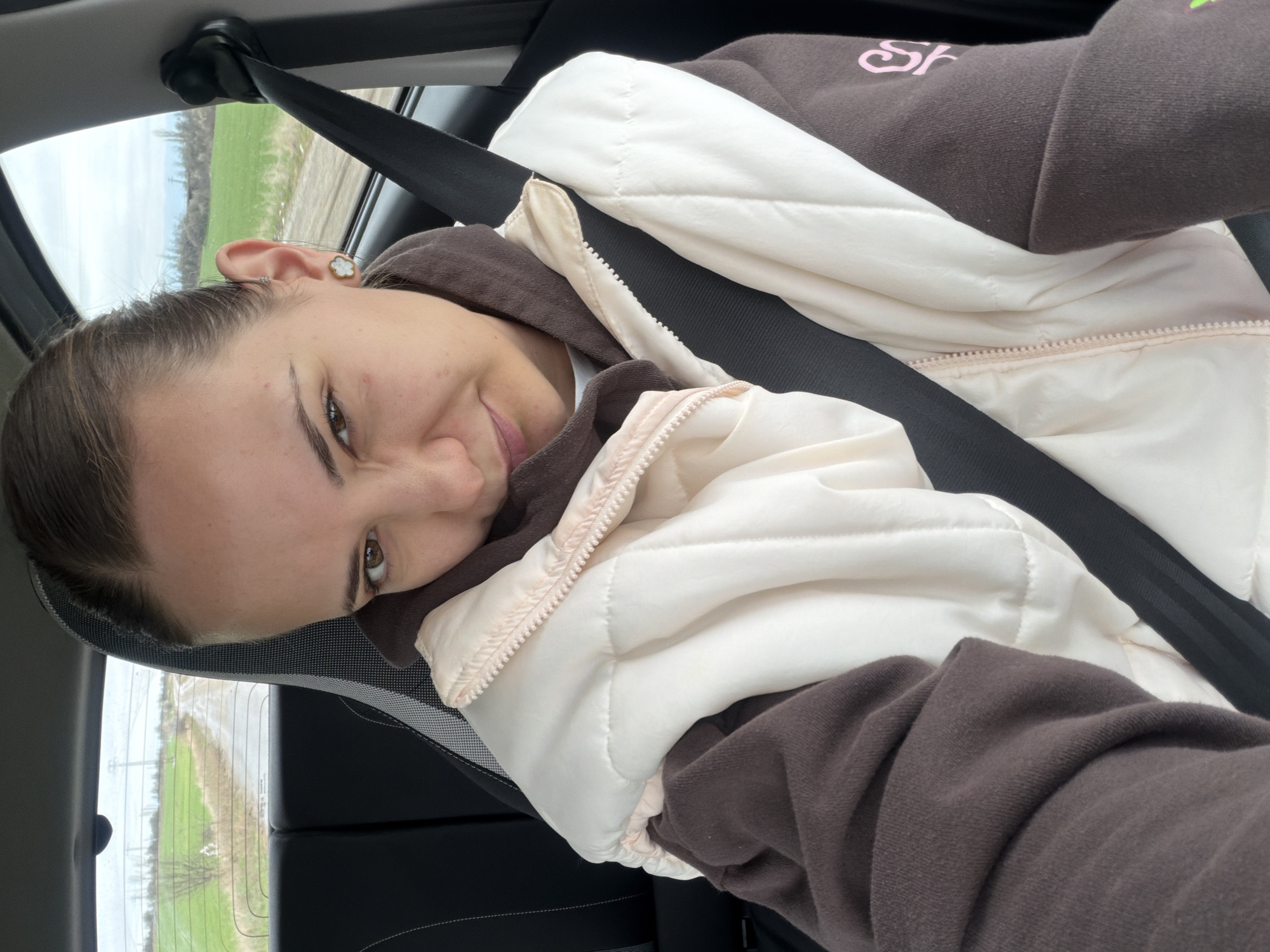 A woman is sitting in a car, wearing a seatbelt and a brown hoodie under a white puffer vest. She is looking at the camera with a slight smile. The car is moving on a road, visible through the window, with fields on either side. The location is not in the city of Sofia, Bulgaria, but rather a rural area.