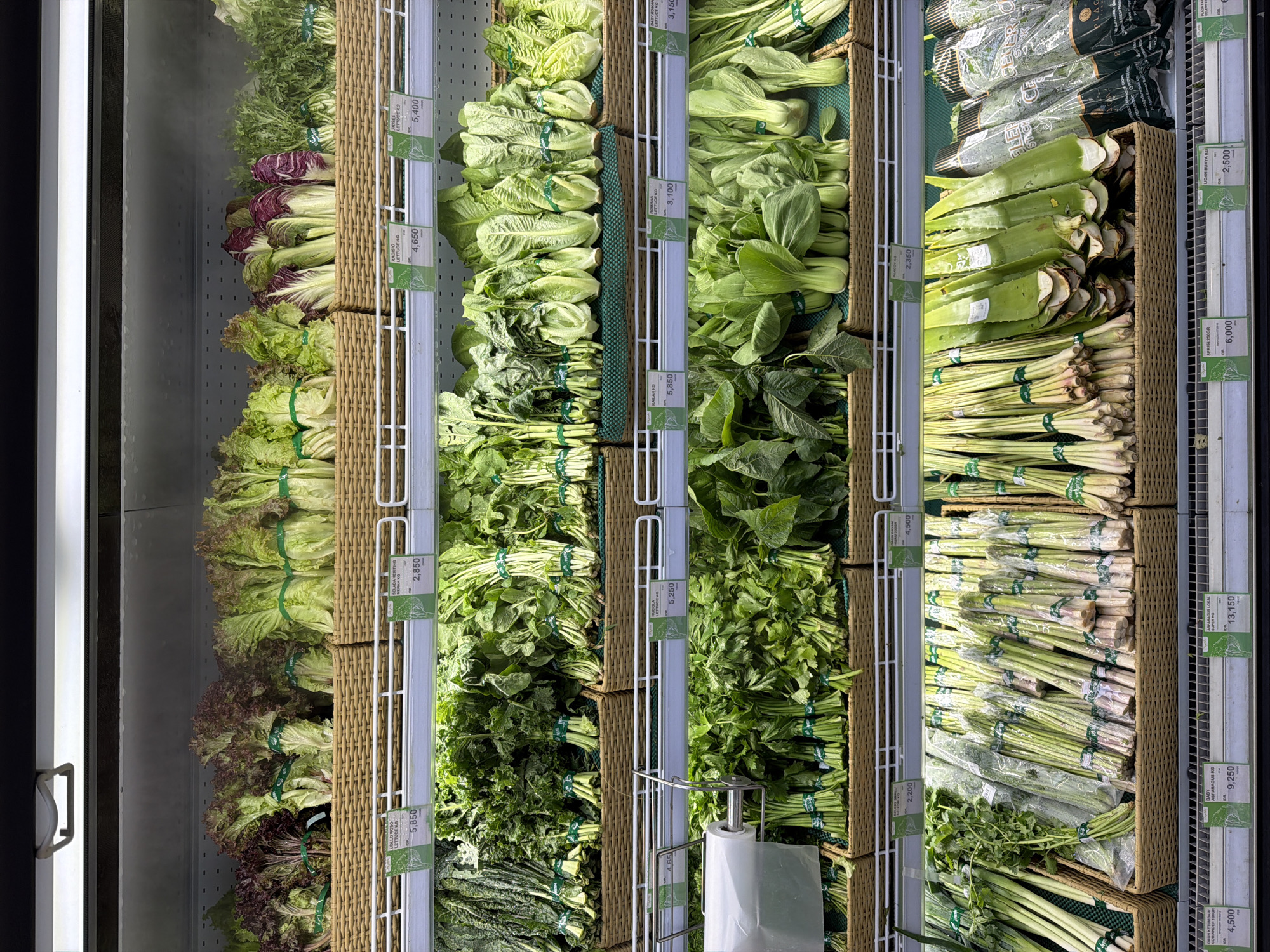 Here is a detailed description of the image:
**Scene:** A display case in what appears to be a grocery store setting.
**Location:** Judging by the Indonesian price tags (IDR) Kedewatan, Indonesia.
**Objects:**
* Various types of leafy green vegetables are arranged on shelves in baskets. Types include (as indicated by price tags): romaine lettuce, baby romaine lettuce, bok choy, kale, radish, butter lettuce, and curly leaf lettuce. Other items include aloe vera and asparagus.
* Each type of vegetable is tied together in bundles with green string.
* Price tags in Indonesian Rupiah (IDR) are attached next to each type of vegetable.
* A roll of plastic bags hangs on a wire frame.
**Environment:**
* The interior lighting is bright.
* The display is inside a refrigerated case with a visible glass door.
* Vertical shelving units are visible.
**Overall Impression:** The image depicts a well-stocked and organized produce section within a grocery store. The various leafy greens are fresh and neatly arranged to attract shoppers.
