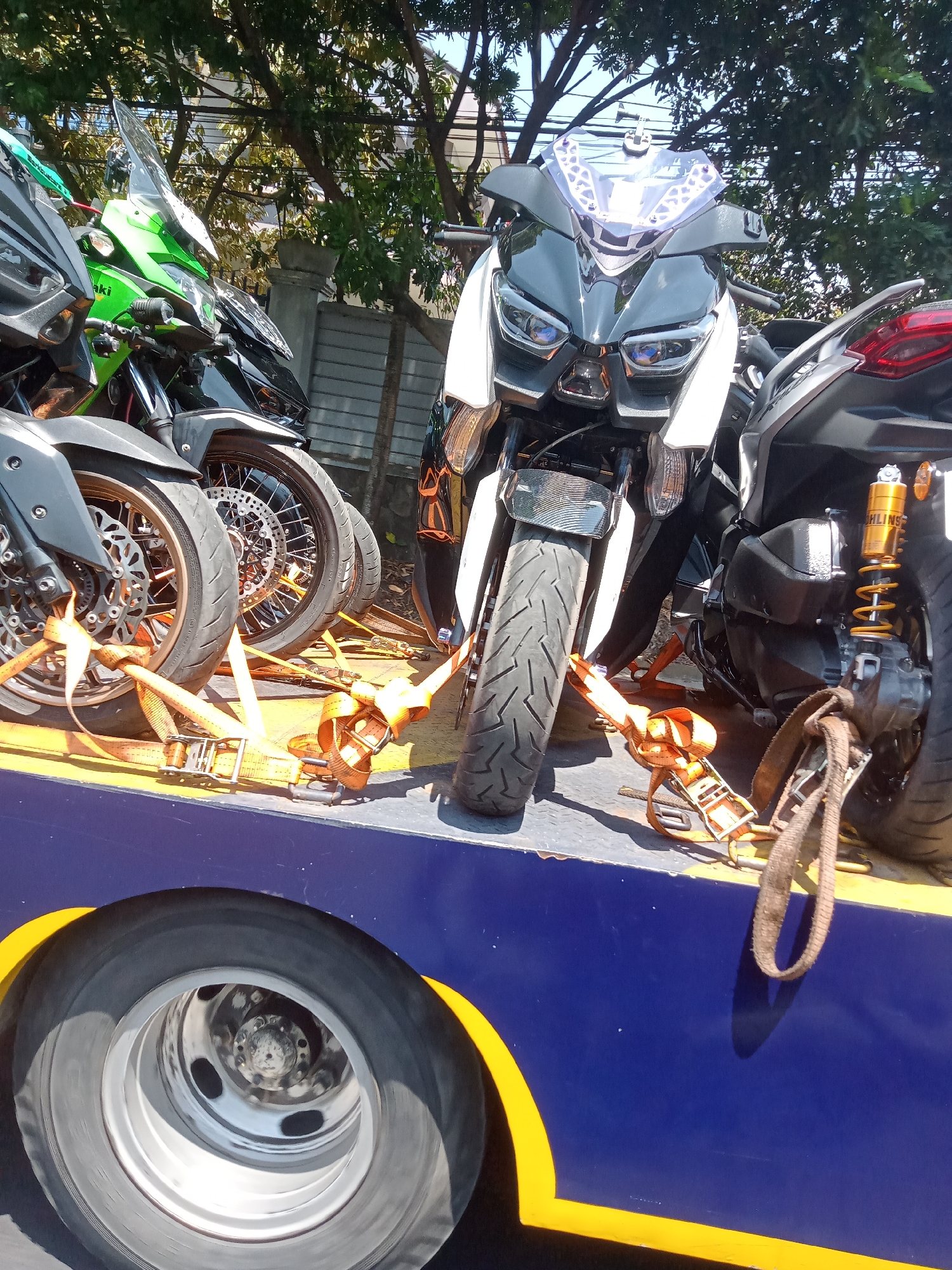 In Batu, Indonesia, on a sunny day, several motorcycles are secured to a flatbed trailer with orange ratchet straps. The motorcycles, a mix of sport bikes and scooters, are positioned tightly together. In the foreground, the large tire and wheel of the trailer are visible. The trailer itself is painted blue with a yellow stripe along the edge. Trees and wires form the background, suggesting an outdoor, possibly roadside, location. No people are visible in the image.