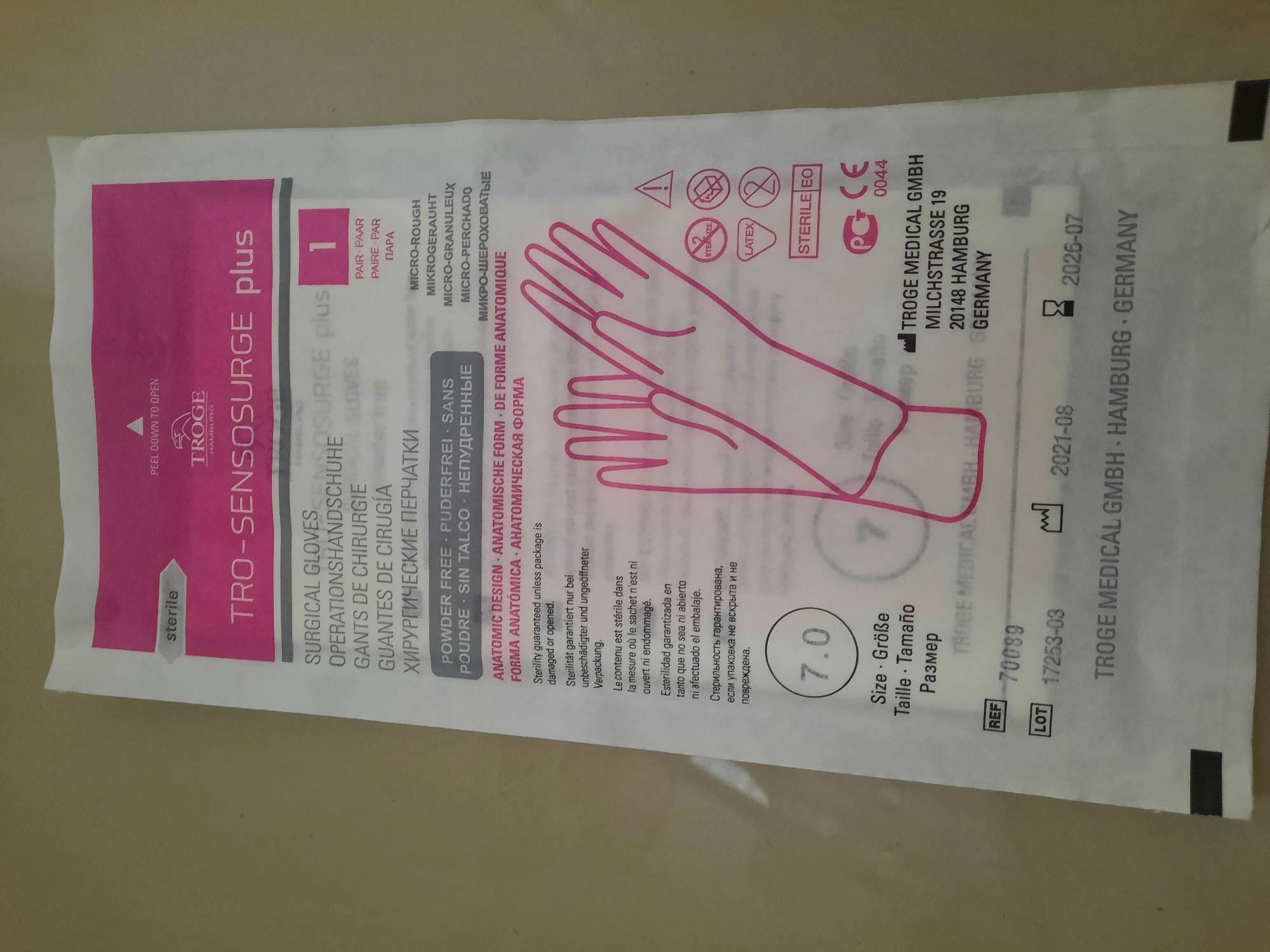 The image is a close-up of a sterile surgical glove package. The package is predominantly white with pink and black lettering and graphics. Text on the package includes "STERILE," "TRO-SENSOSURGE plus," "SURGICAL GLOVES," and multilingual descriptions of the product. There is also a size indication of "7.0." A graphic representation of a gloved hand is visible. Below this, there are various symbols including "STERILE EO" and a CE mark. Manufacturer details are printed at the bottom: "TROGE MEDICAL GMBH MILCHSTRASSE 19 20148 HAMBURG GERMANY." There are also lot and reference numbers, along with manufacturing and expiry dates. The background of the image is a plain, light-colored surface, consistent with a table or floor. The image does not contain any elements that would indicate its location in Lhokseumawe, Indonesia.