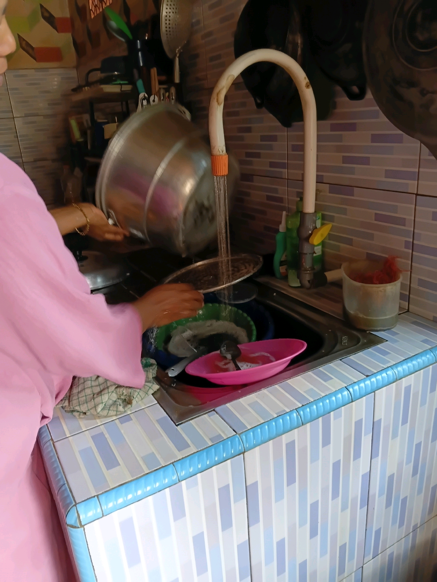 In a kitchen in Margo Rukun, Indonesia, a person wearing a pink garment is washing dishes. The sink area is tiled with blue and white patterns, and a faucet is running water into the sink. There are several dishes in the sink, including a large metal pot and a pink bowl. On the counter, there are cleaning supplies and a container with red bristles. The background shows shelves with kitchenware.